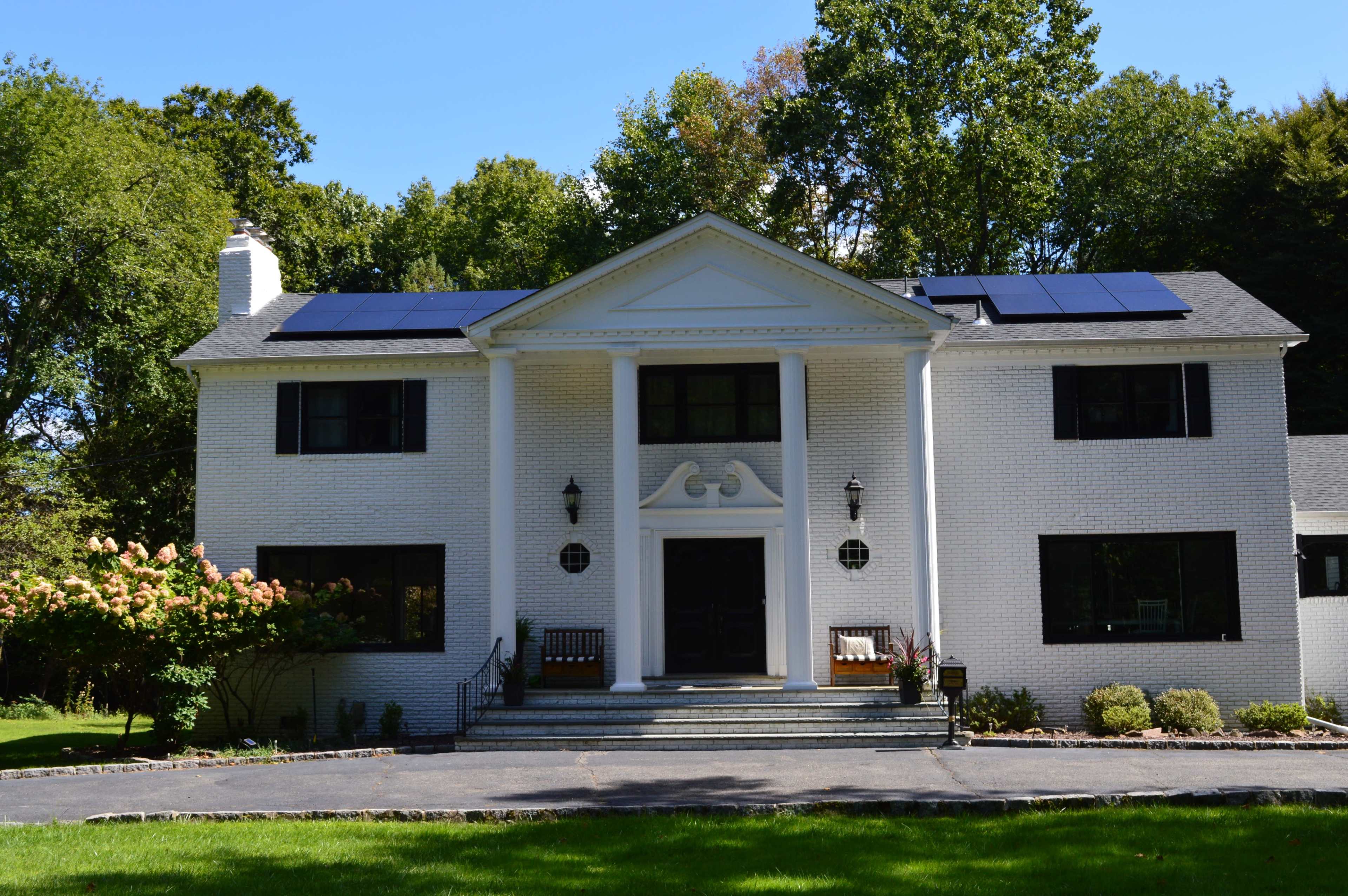 The image shows a two-story white brick house with a front stairway, columns, solar panels on the roof, and a well-manicured lawn.