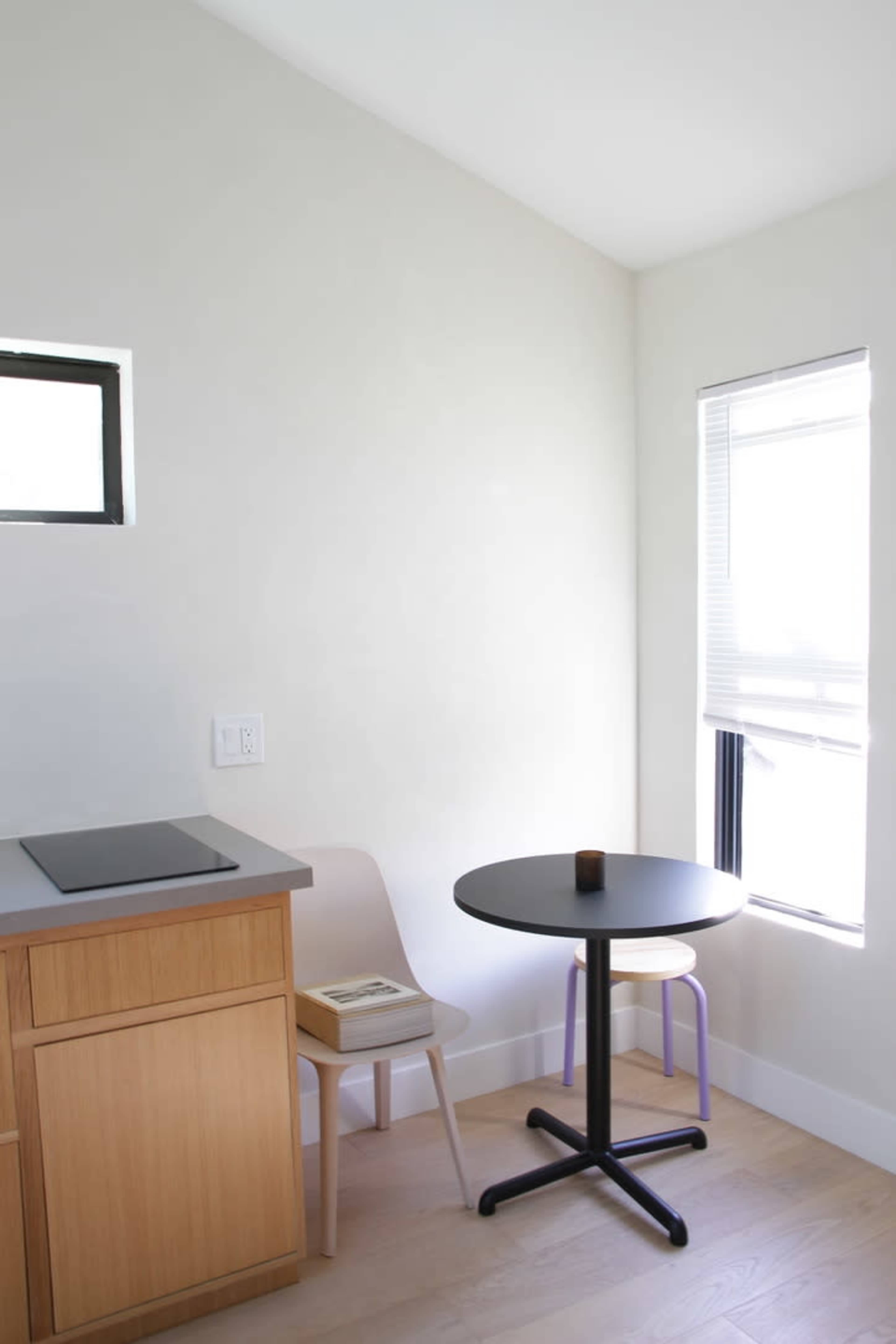 A small corner kitchen area featuring a table with a black top and two chairs, one beige and one purple, beside a window with blinds.