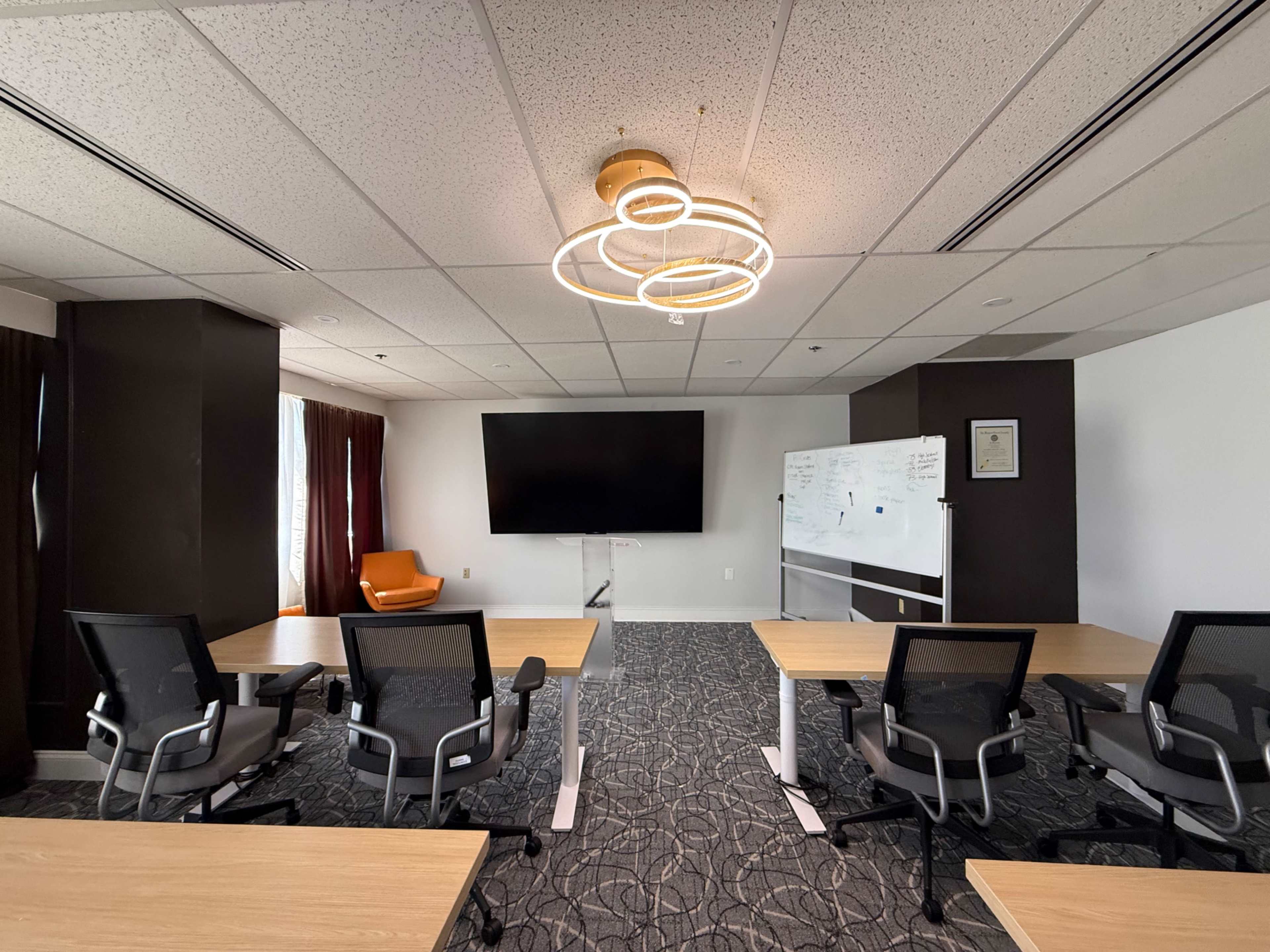 The image shows a modern conference room with multiple tables, chairs, a large screen on the wall, and a whiteboard.