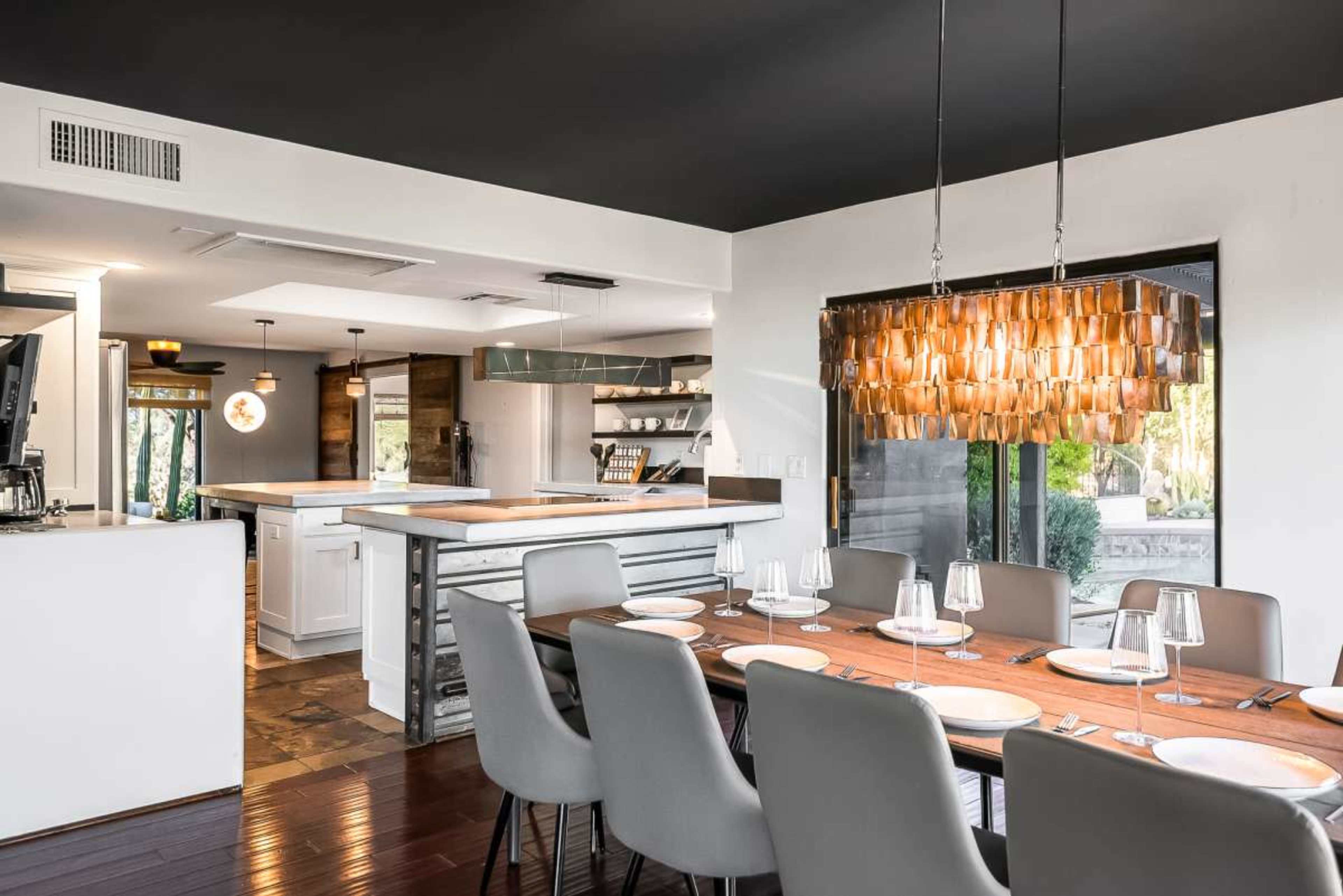 The image features a contemporary kitchen and dining area with a wooden dining table set for a meal, surrounded by modern chairs and highlighted by a distinctive pendant light overhead.