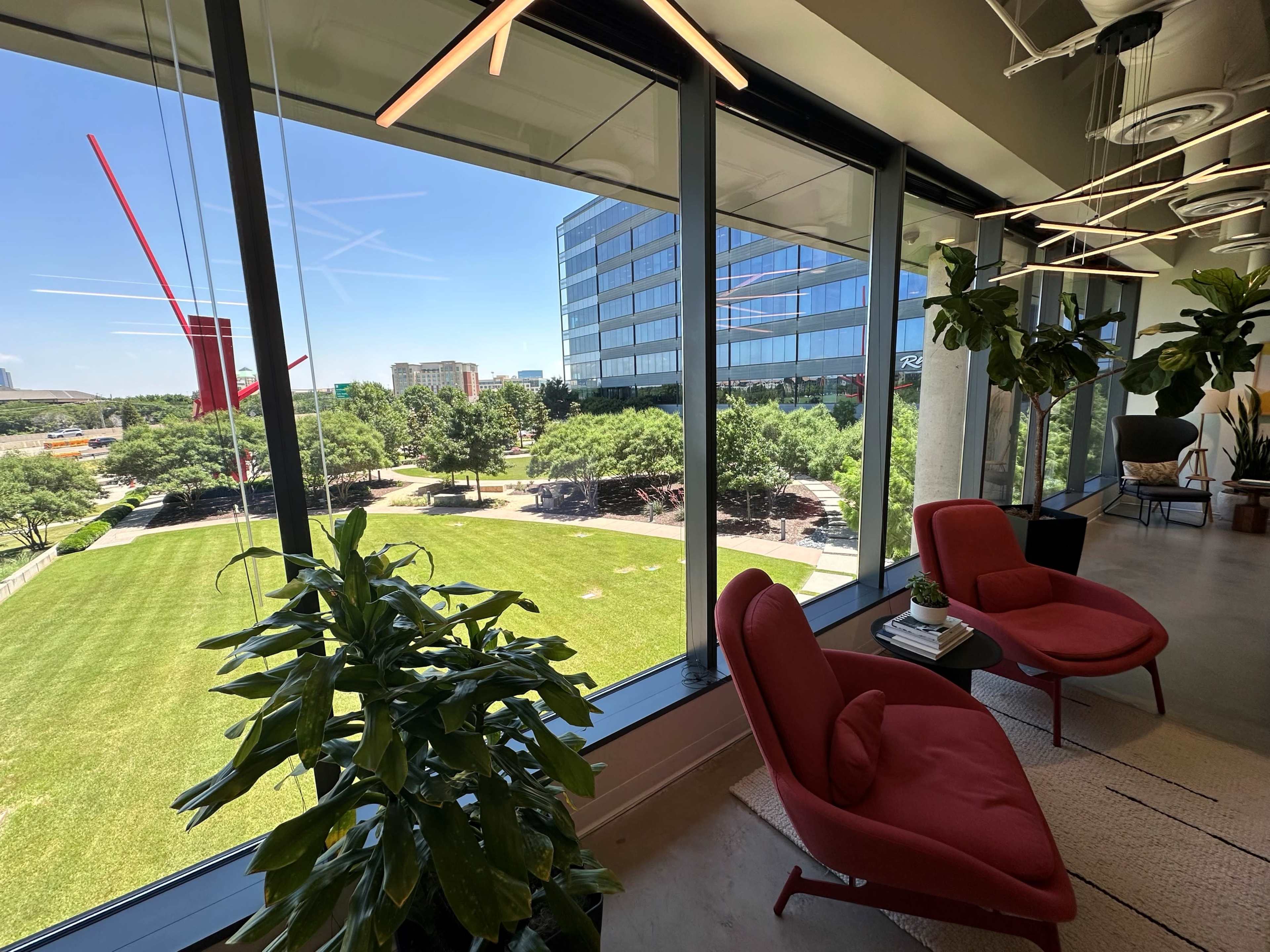 A modern office space with two red chairs, large windows overlooking a green park, and plants in the foreground.