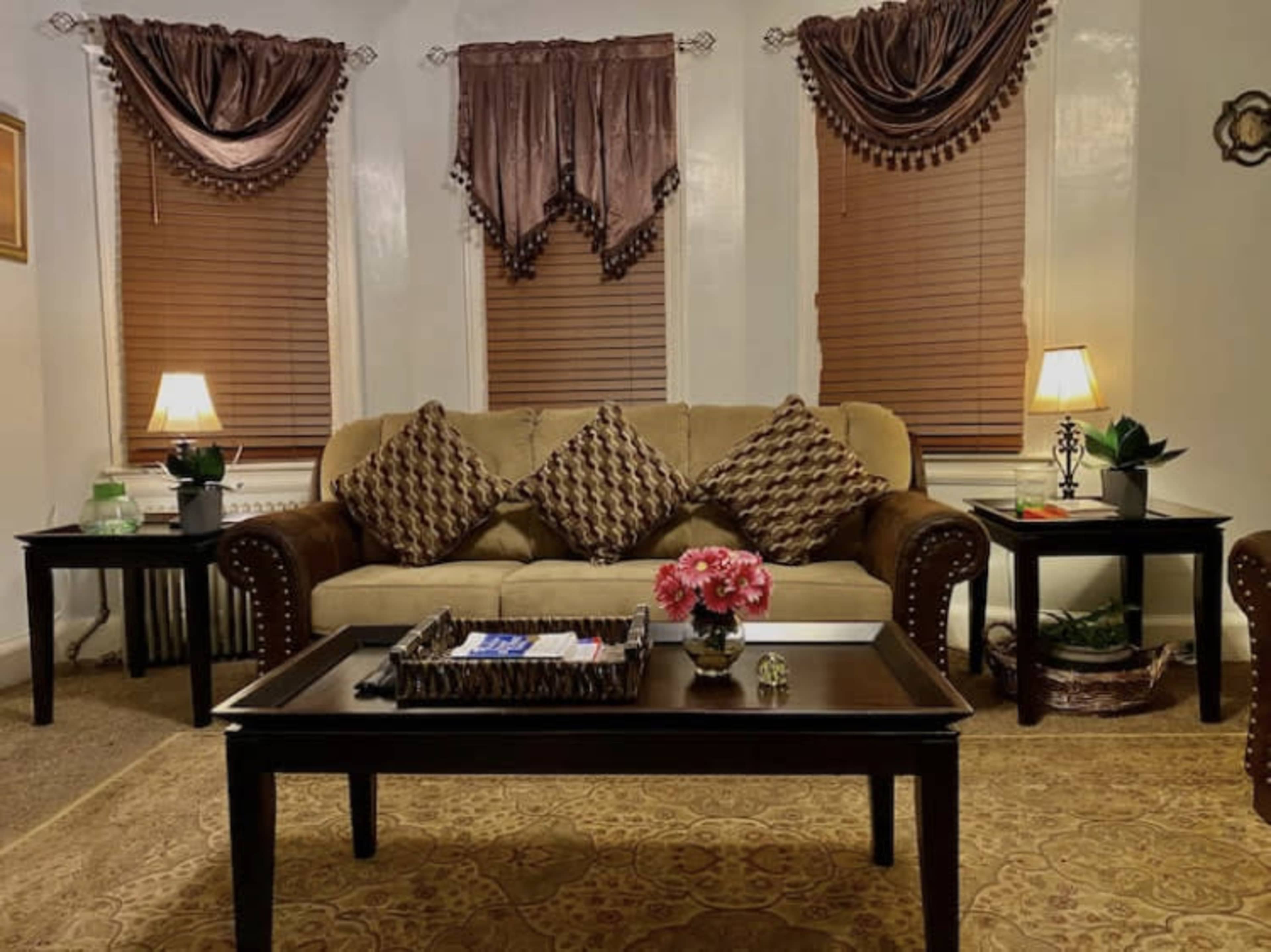 The scene depicts a living room with a beige sofa adorned with decorative cushions, accented by two tables with lamps and plants, all set against a backdrop of brown curtains and a patterned rug.