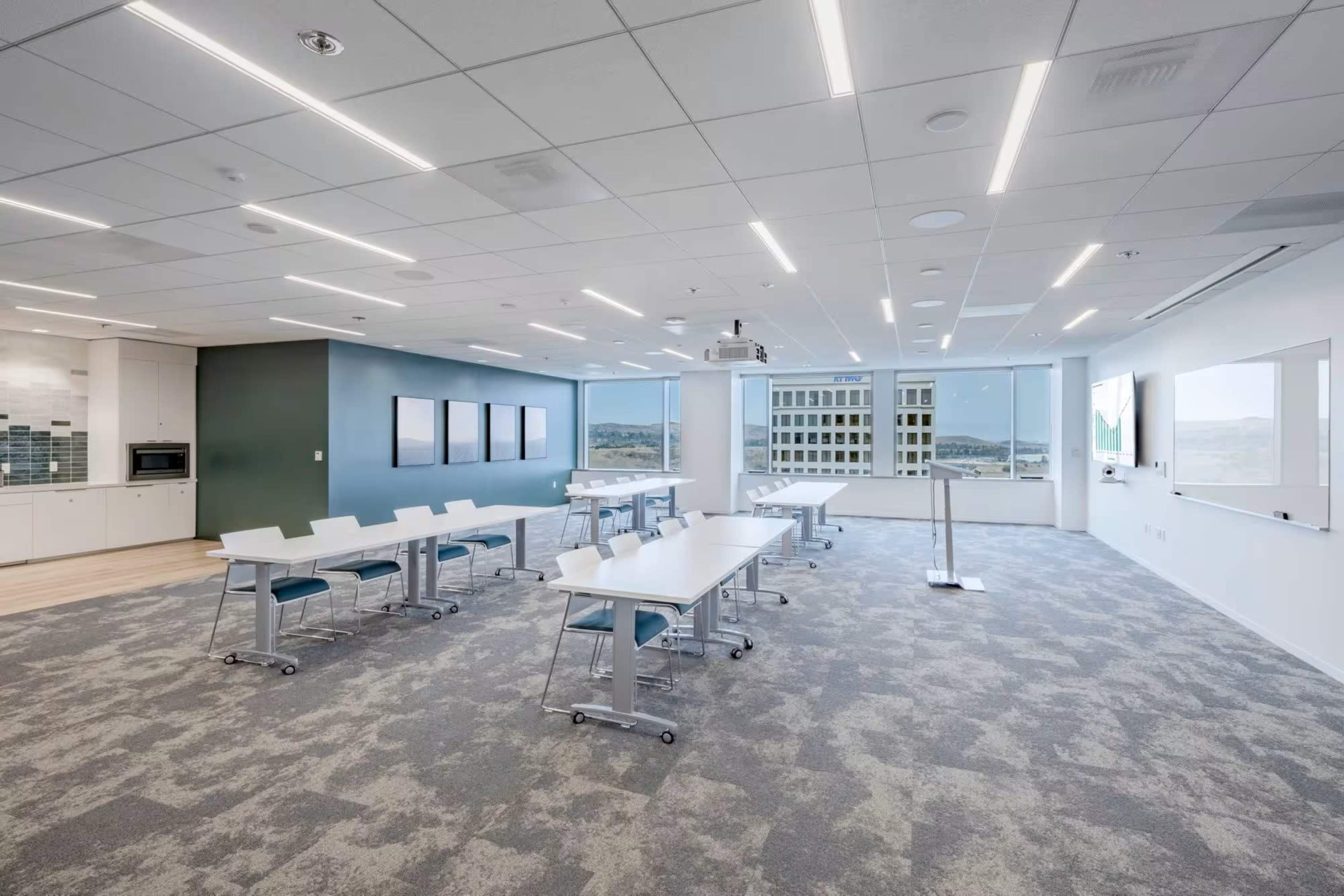 The image shows a modern conference room with several white tables, chairs, and large windows allowing natural light to enter.
