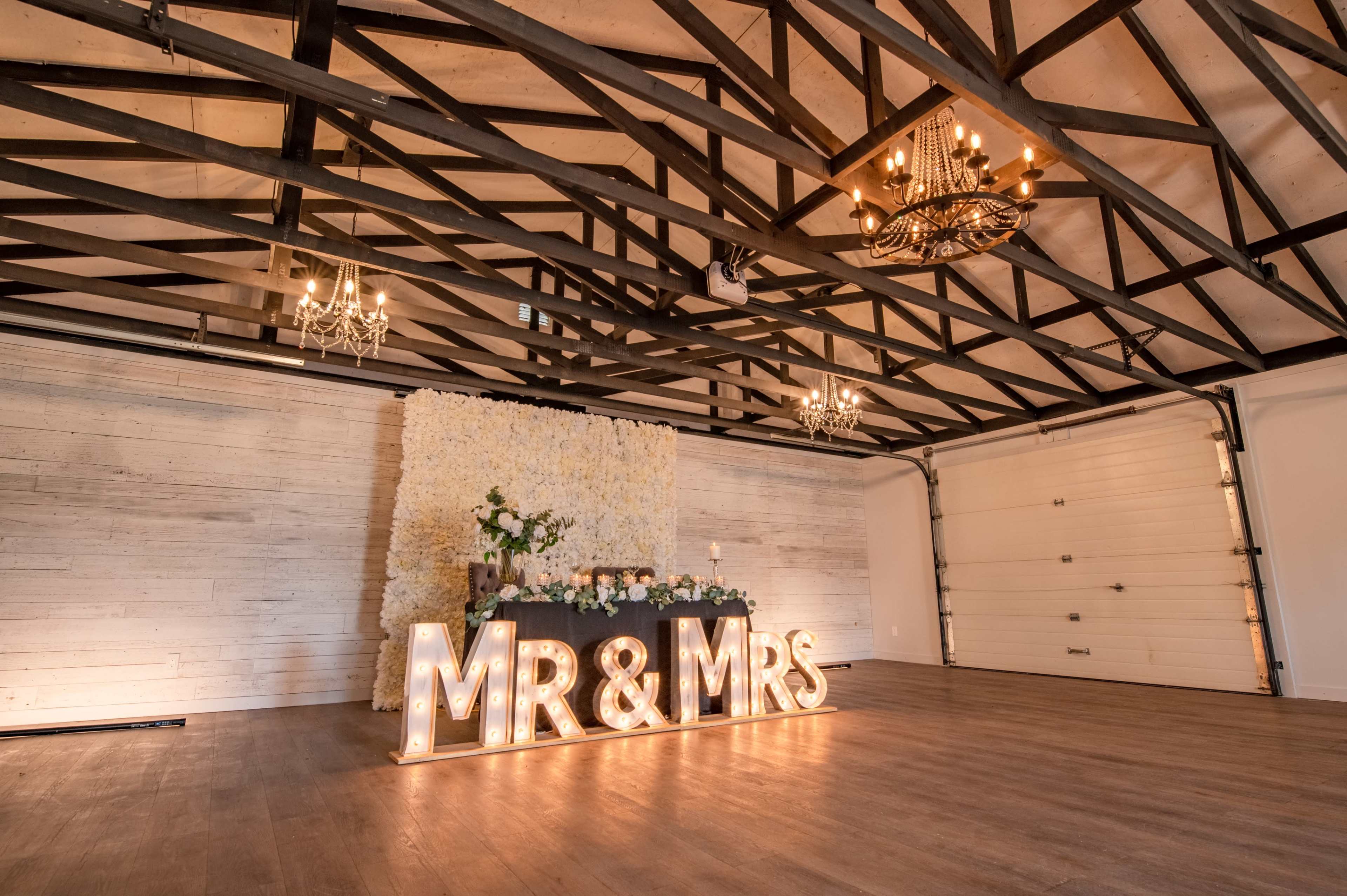 The image shows a decorated indoor space featuring a large illuminated "MR & MRS" sign in front of a floral backdrop and a small table with candles and decorations.