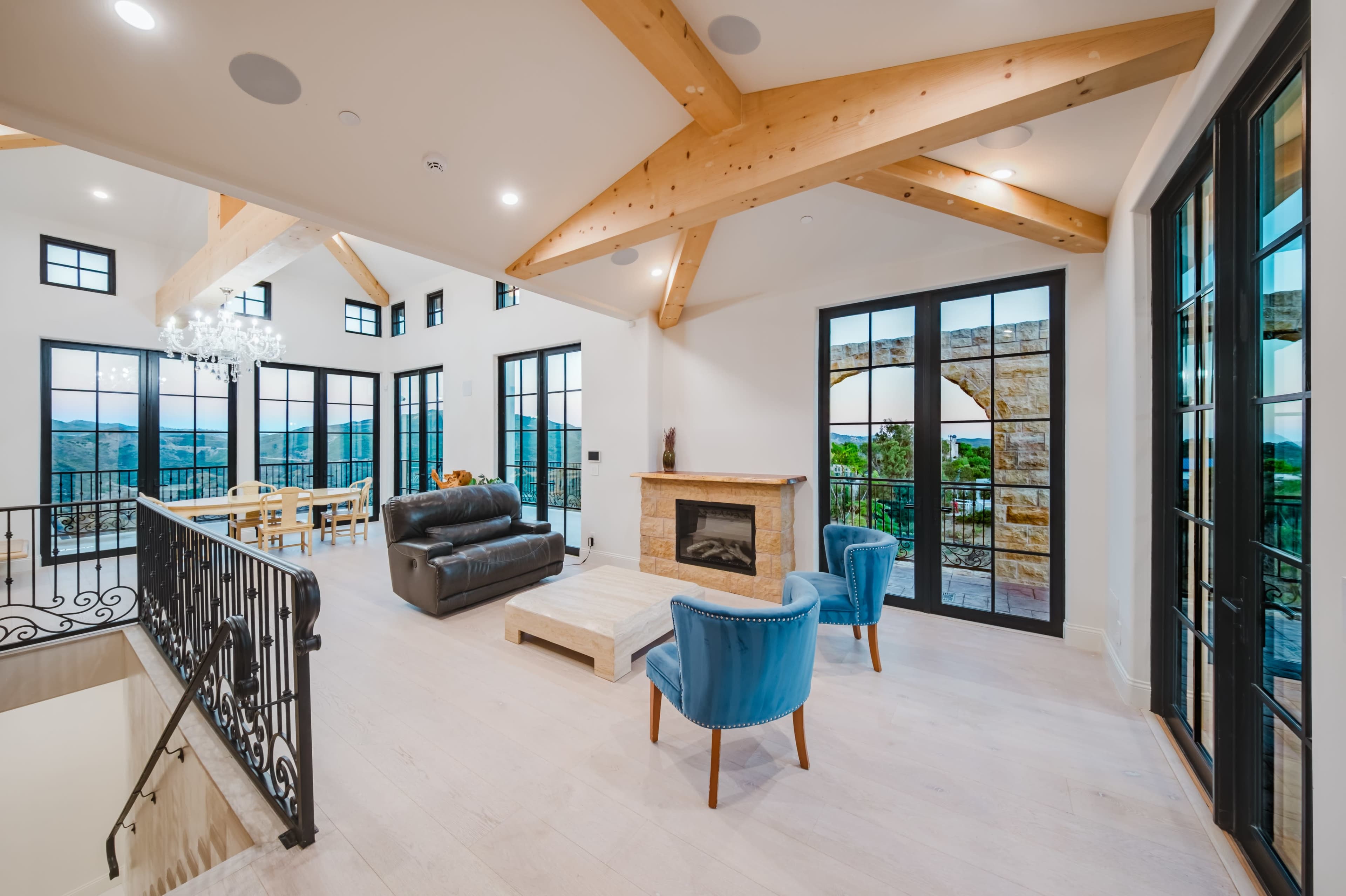 The image shows a spacious living room with large windows, wooden beams, and modern furniture, including a black leather sofa and blue chairs, overlooking a scenic view.