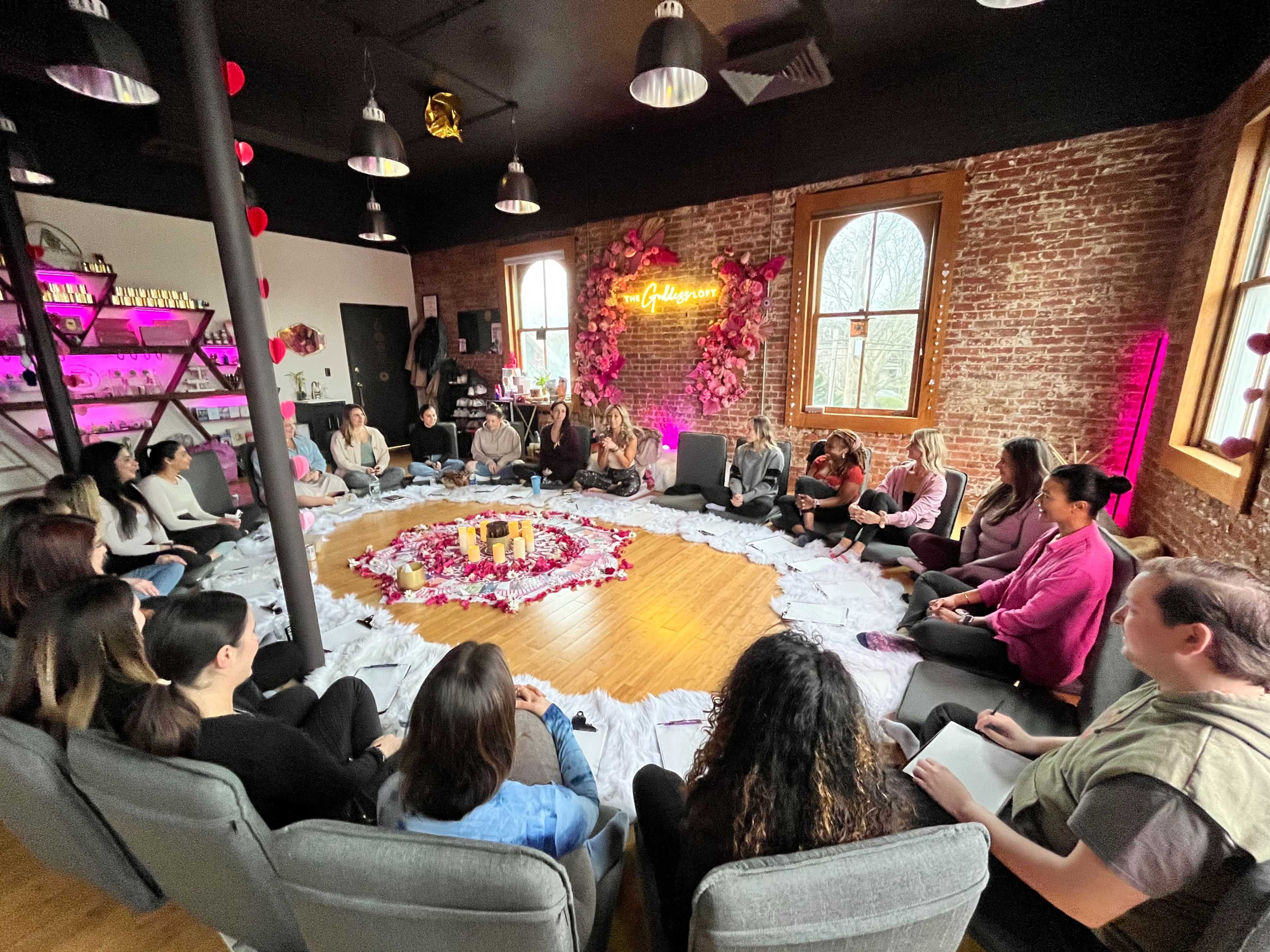 A group of people sits in a circular arrangement on the floor of a brick-walled room, surrounded by decorative flowers and soft lighting.