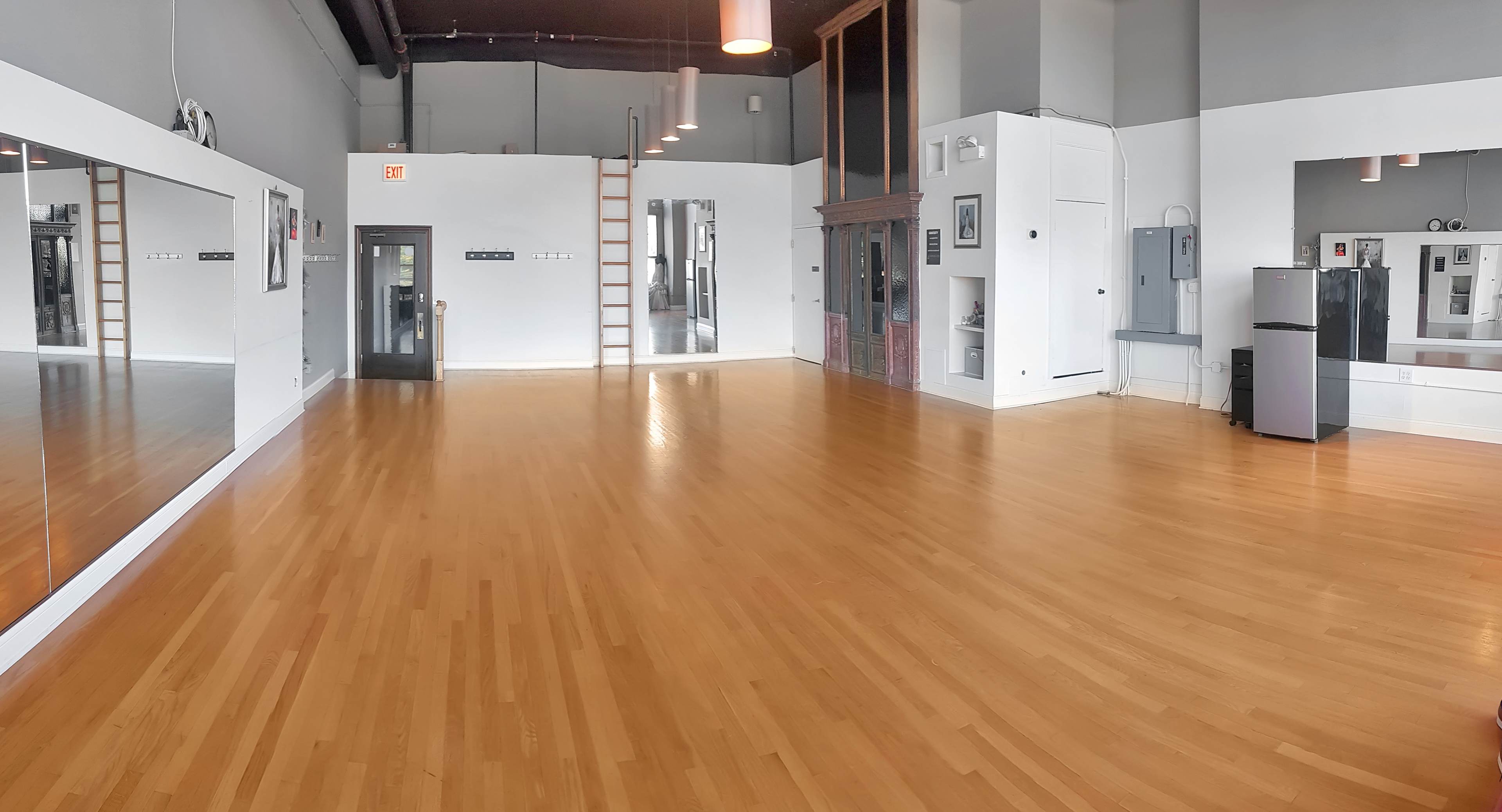 A spacious, empty dance studio with wooden flooring, large mirrors on one wall, and minimal furnishings.