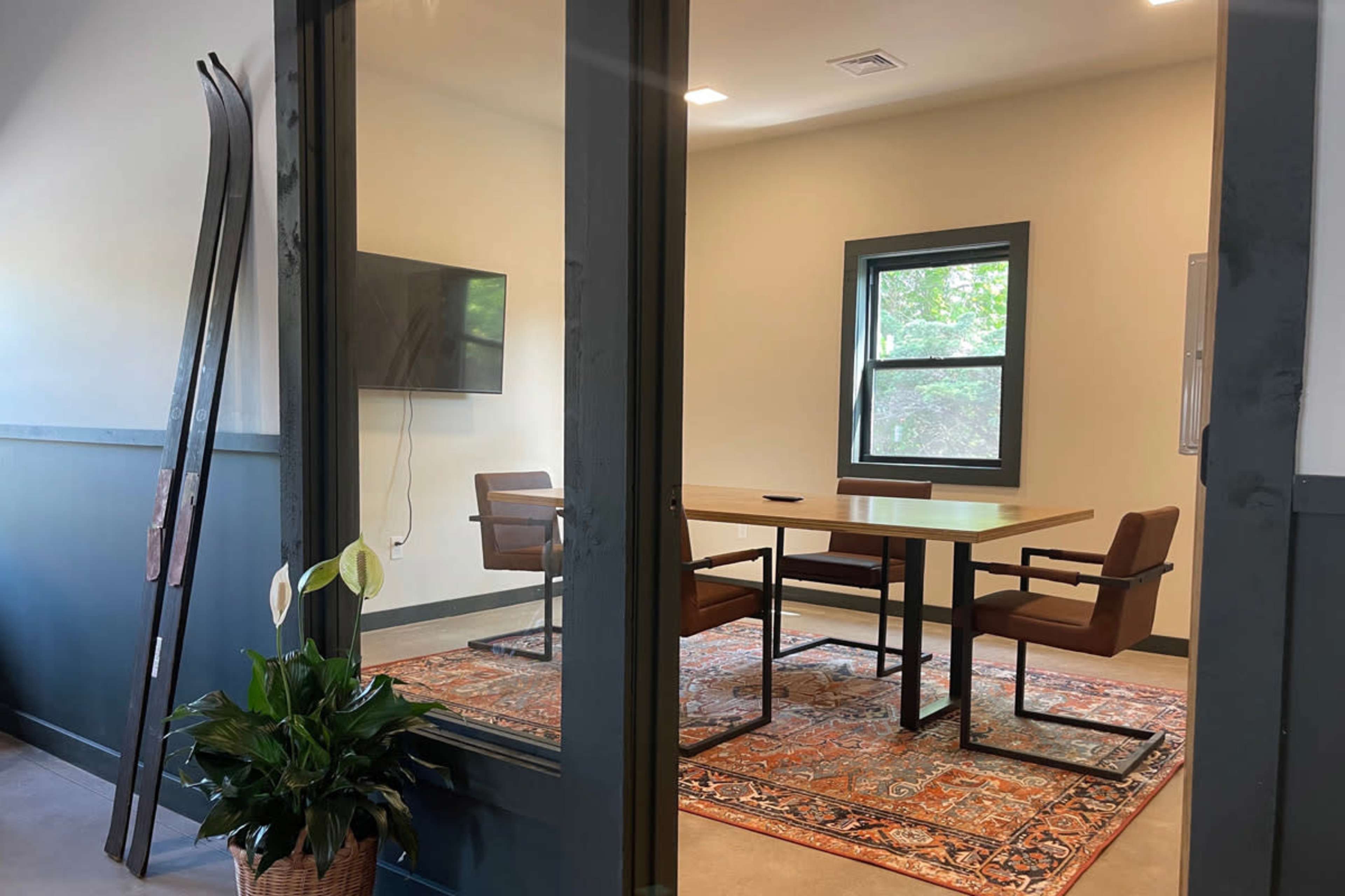 A meeting room features a table with four chairs, a large television on the wall, and a decorative rug, framed by glass doors and a potted plant nearby.