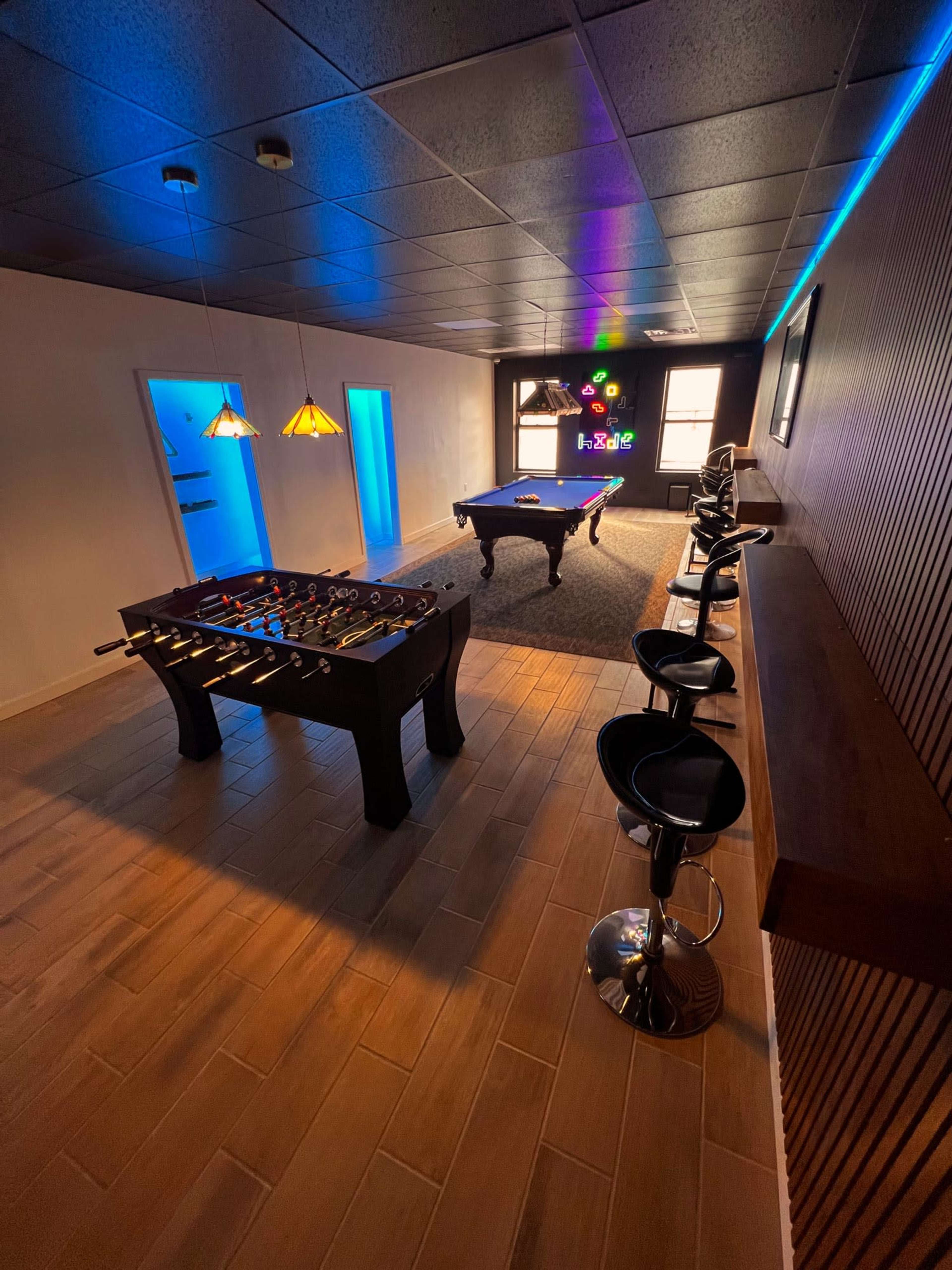 The image shows an entertainment room featuring a foosball table, a billiards table, and bar stools arranged around a counter, with ambient lighting and wooden wall accents.