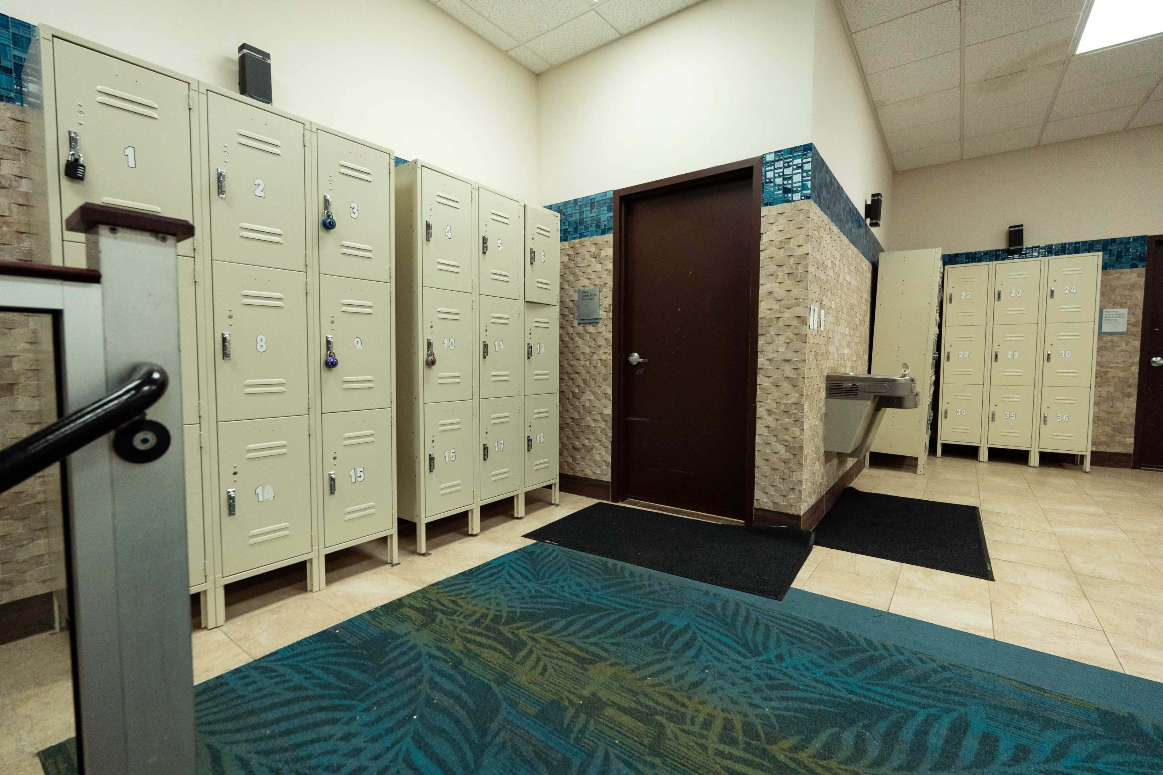 The image shows a room with rows of metal lockers on the left and a door on the right, with a water fountain nearby.