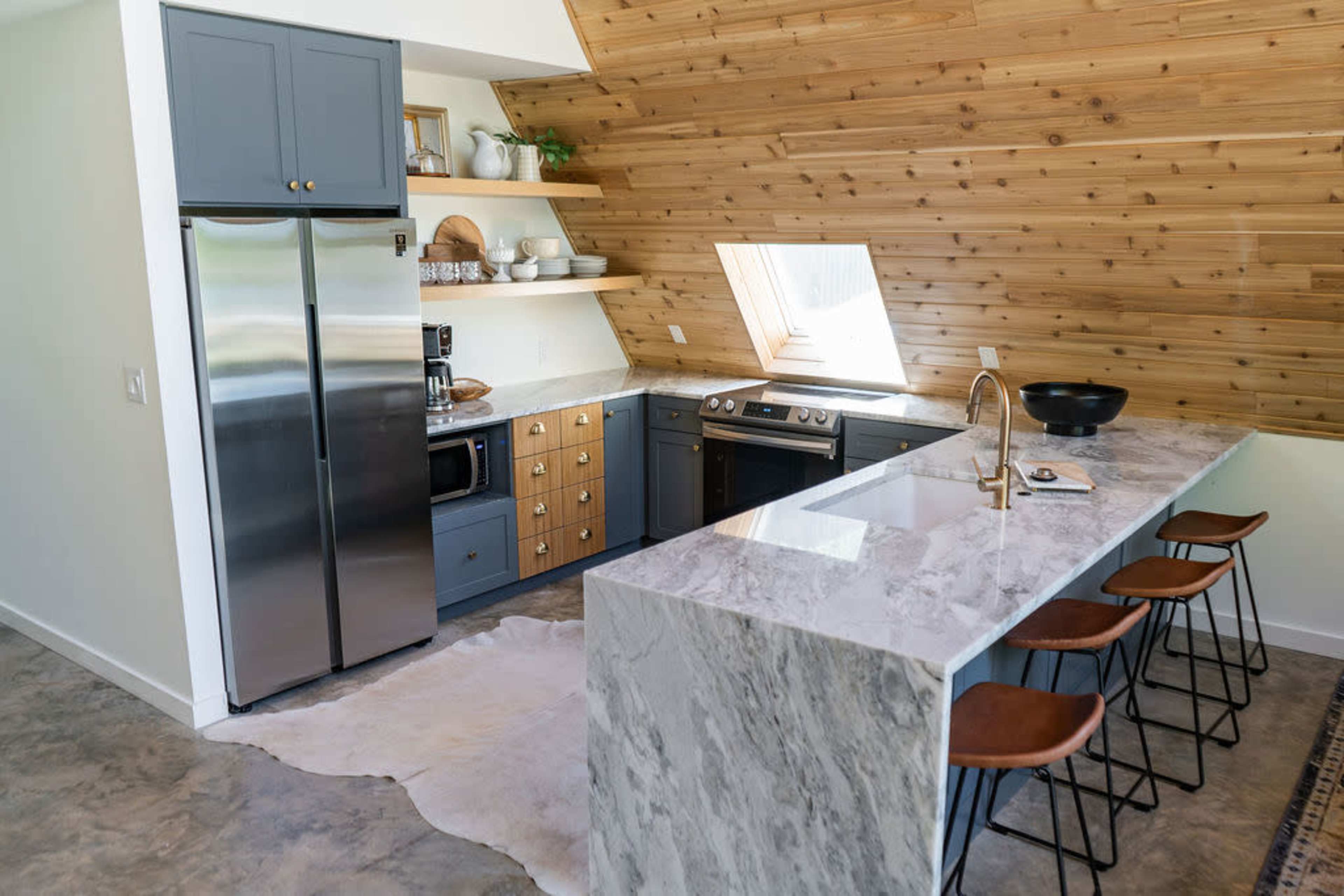 A modern kitchen with gray cabinets, a marble countertop, and wooden walls, featuring an island with four bar stools and a window letting in natural light.