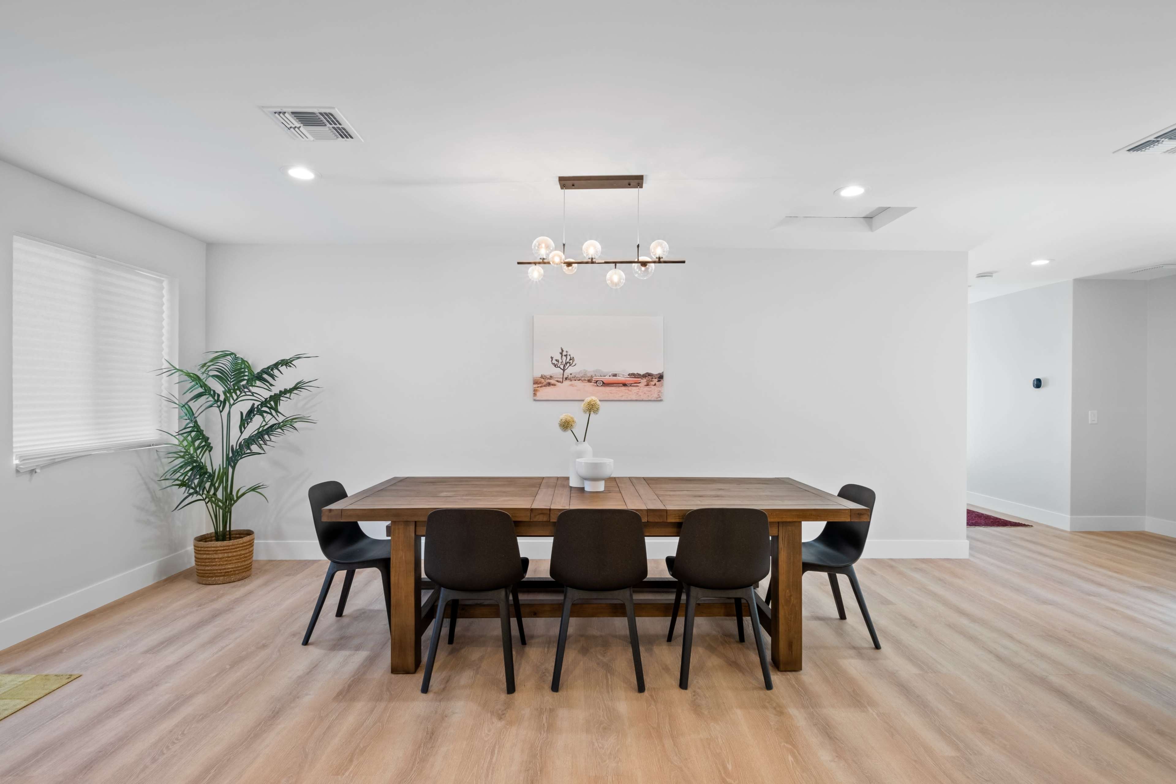 A wooden dining table surrounded by black chairs is positioned in a bright, modern dining area with a potted plant and a piece of artwork on the wall.