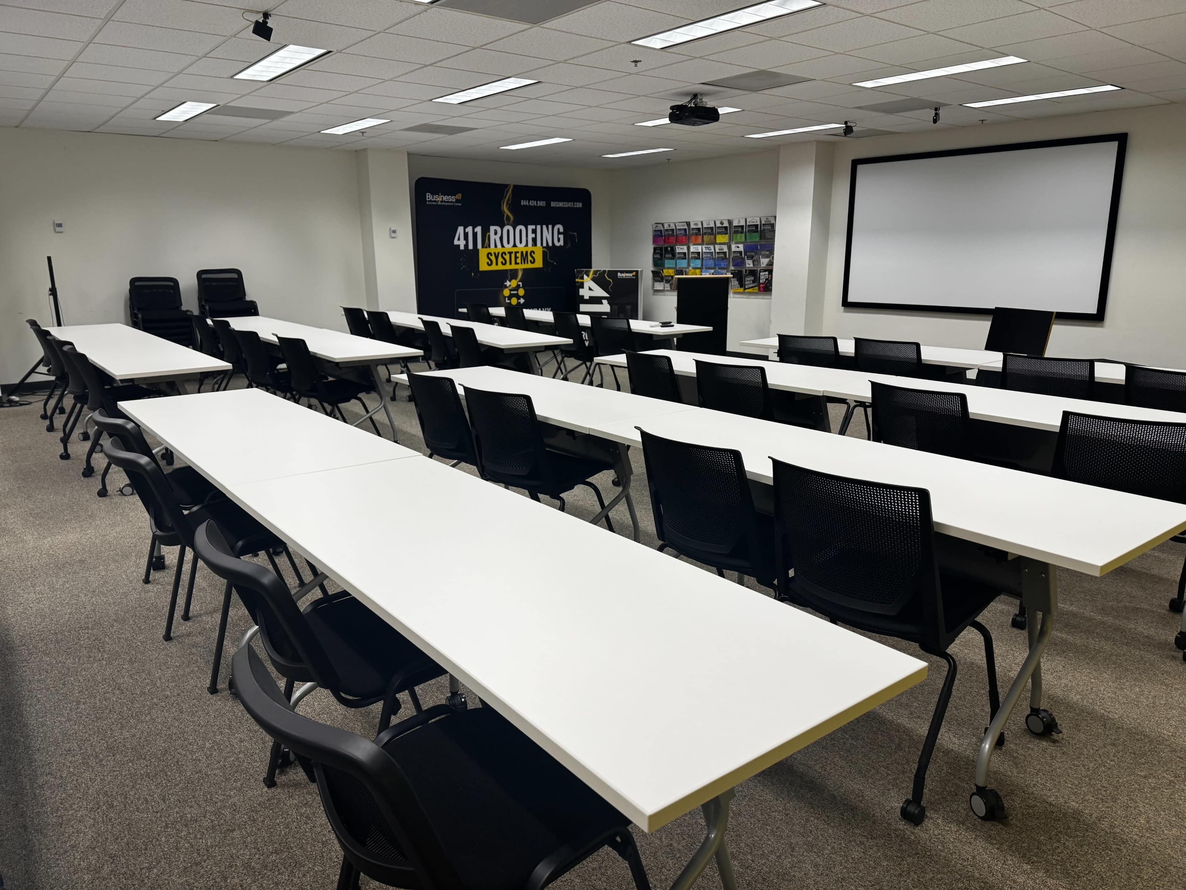 The image shows a training room with multiple long tables arranged in rows, black chairs, and a large screen at one end.