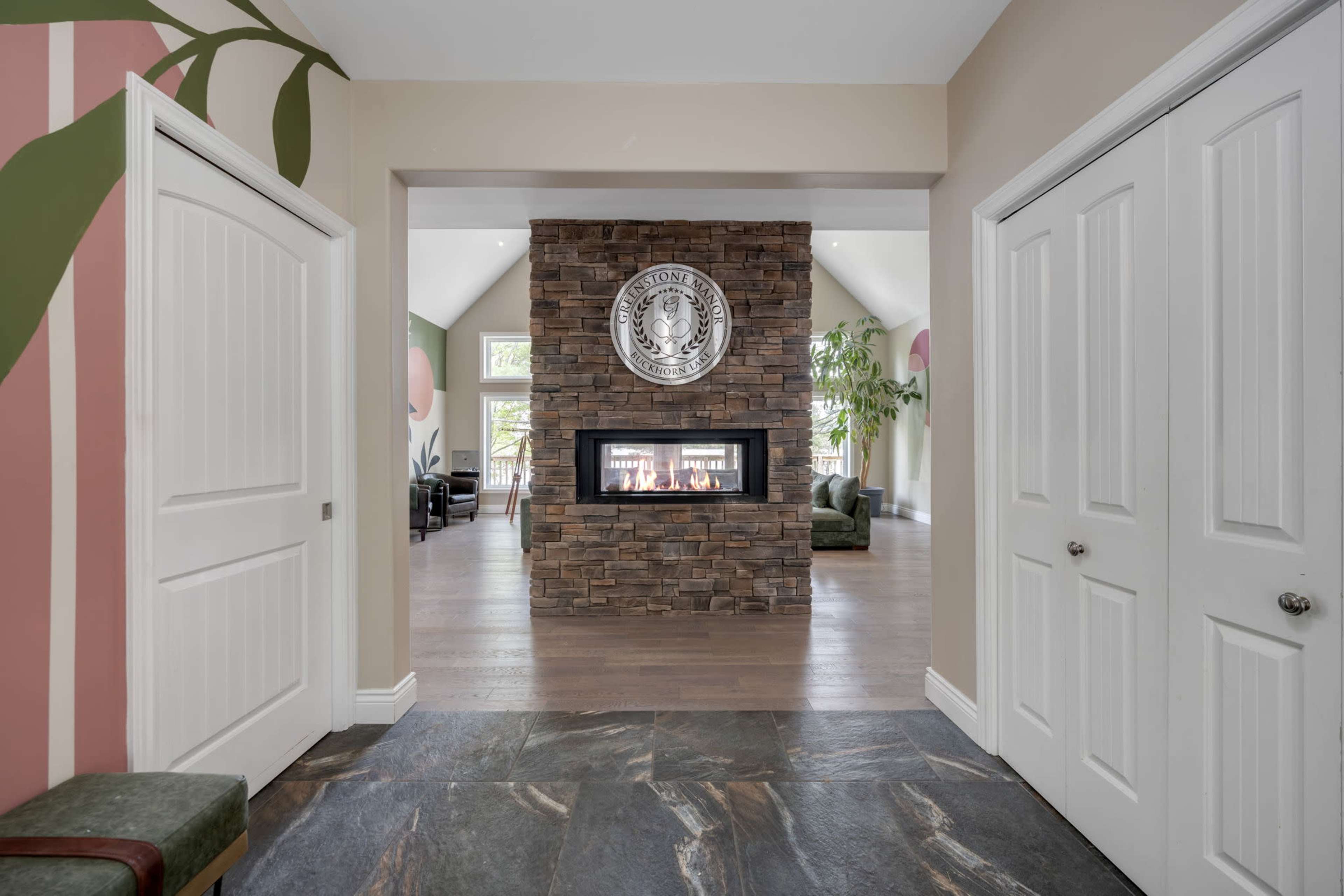 A spacious foyer with a stone fireplace at the center, flanked by two white doors and leading to a living area with green walls and large windows.
