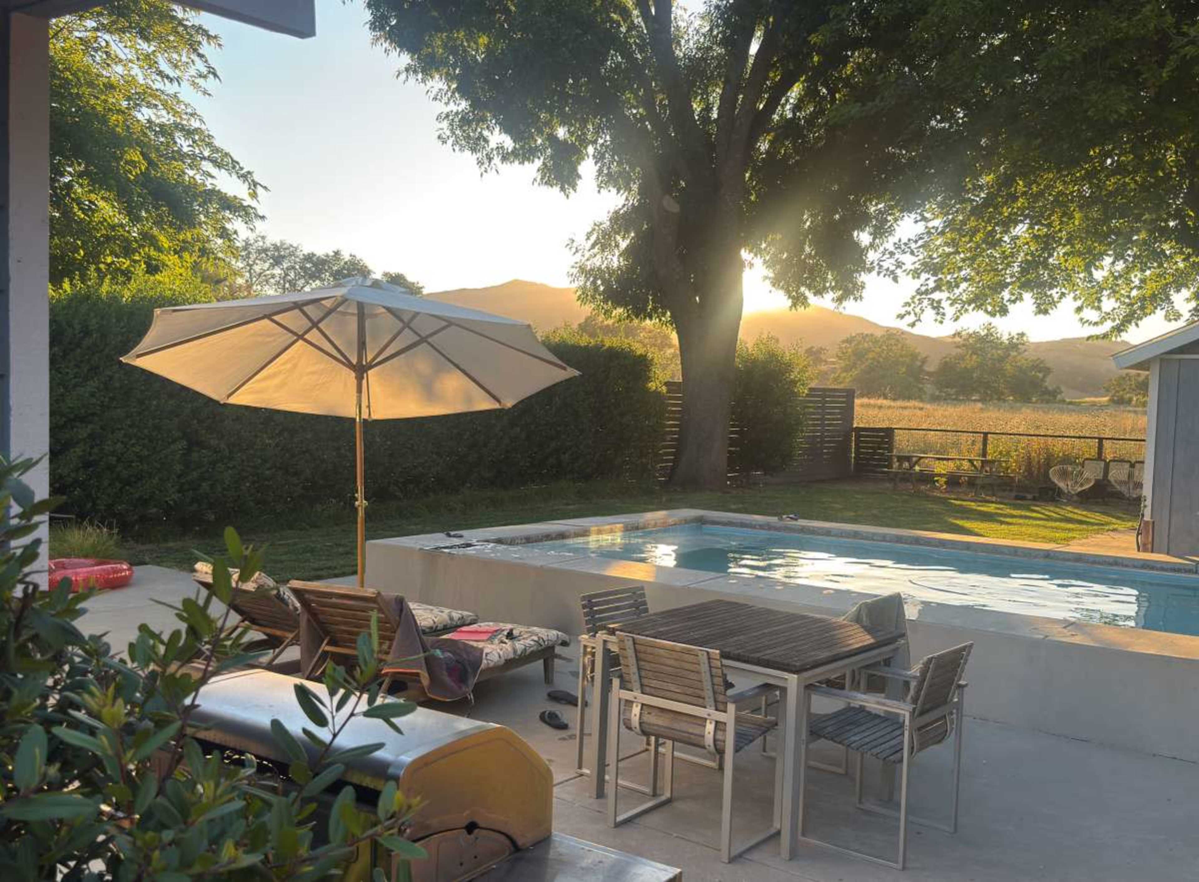 The image features a pool area with a table and chairs under a large umbrella, surrounded by greenery and mountains in the background during sunset.