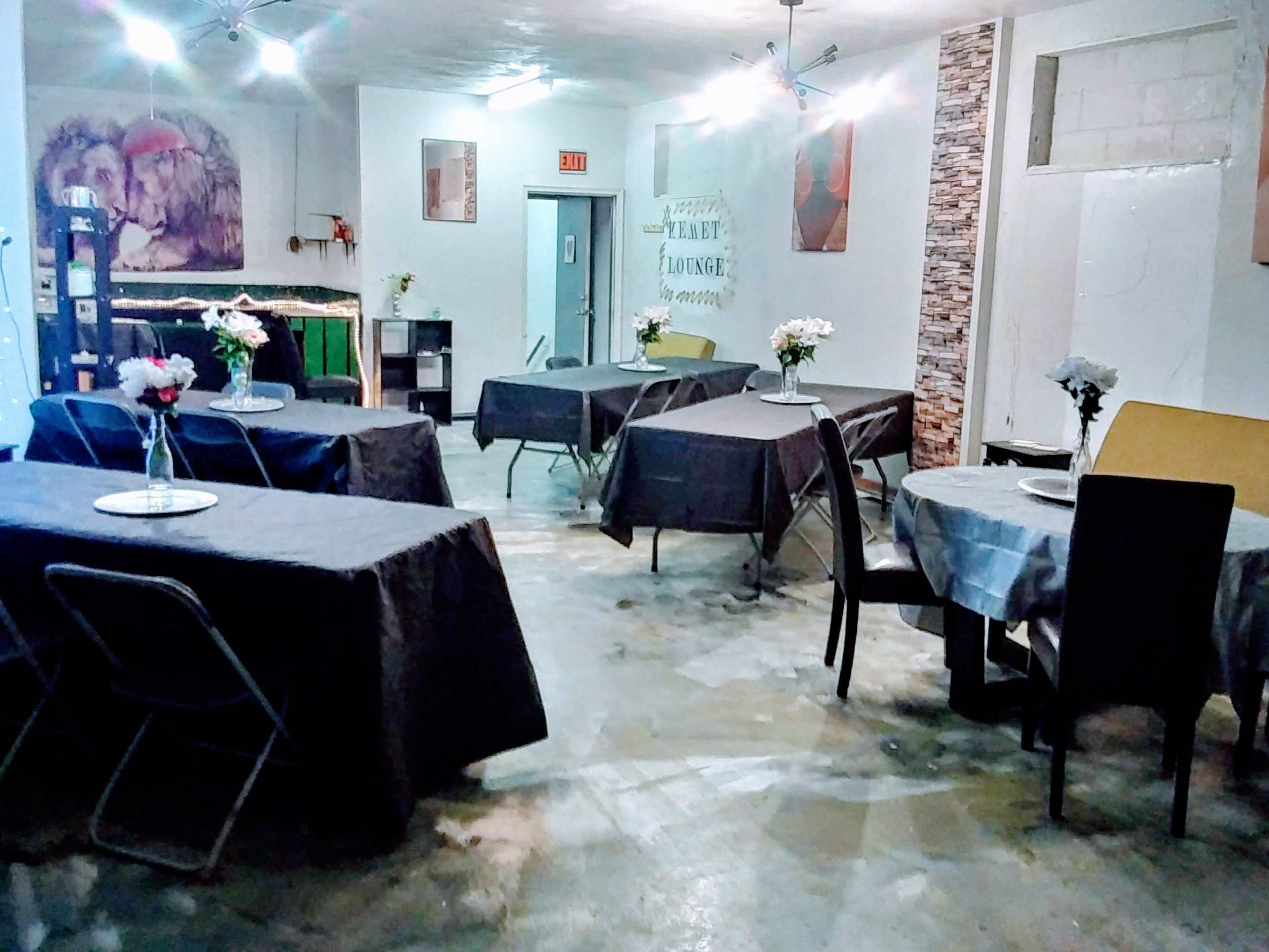 The image shows a dining area with several tables covered in gray tablecloths, arranged in a spacious room with simple decor and flowers on each table.