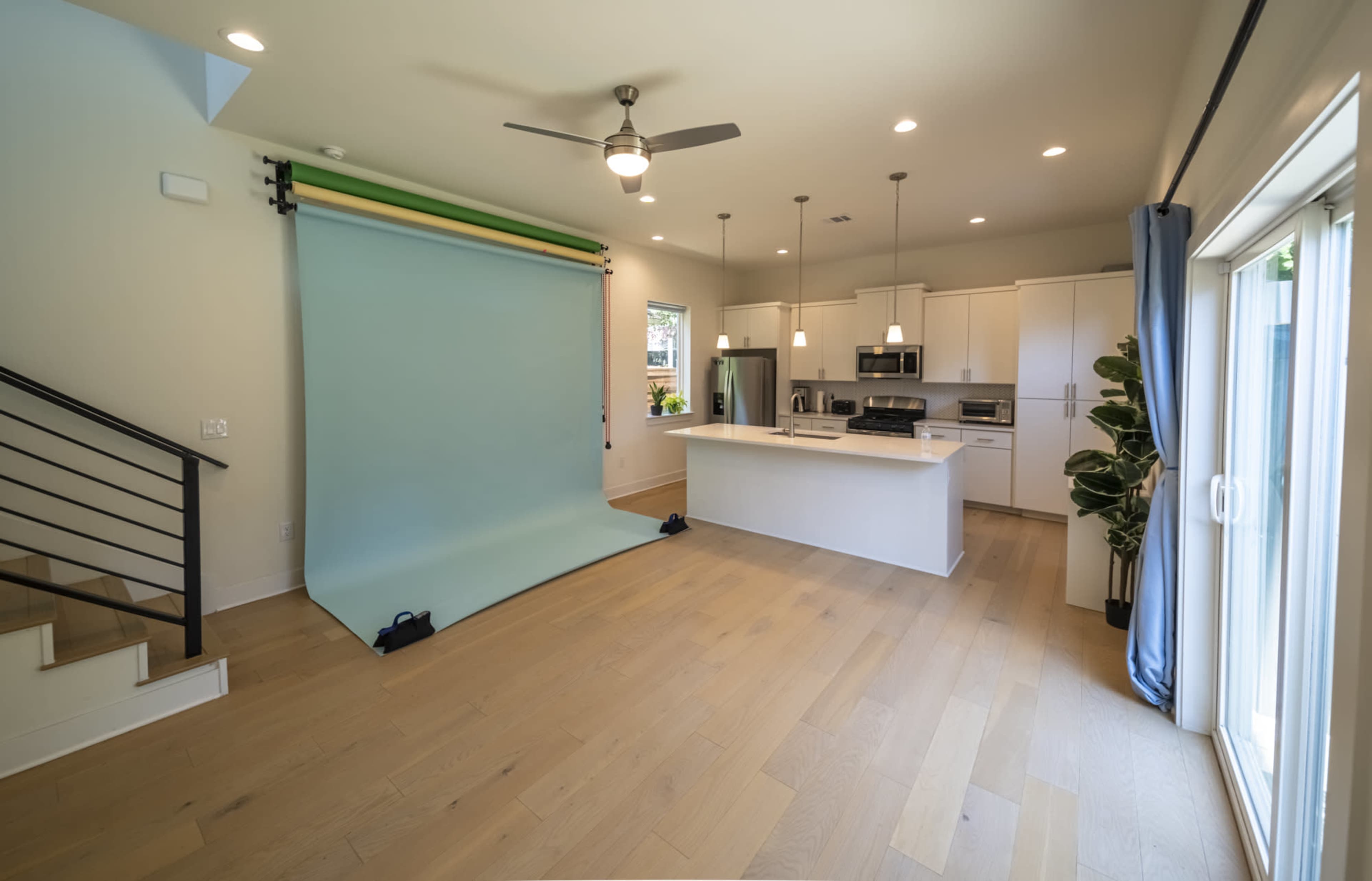 The image shows a spacious room with a green backdrop set up for photography, a kitchen area visible in the background, and a large window letting in natural light.