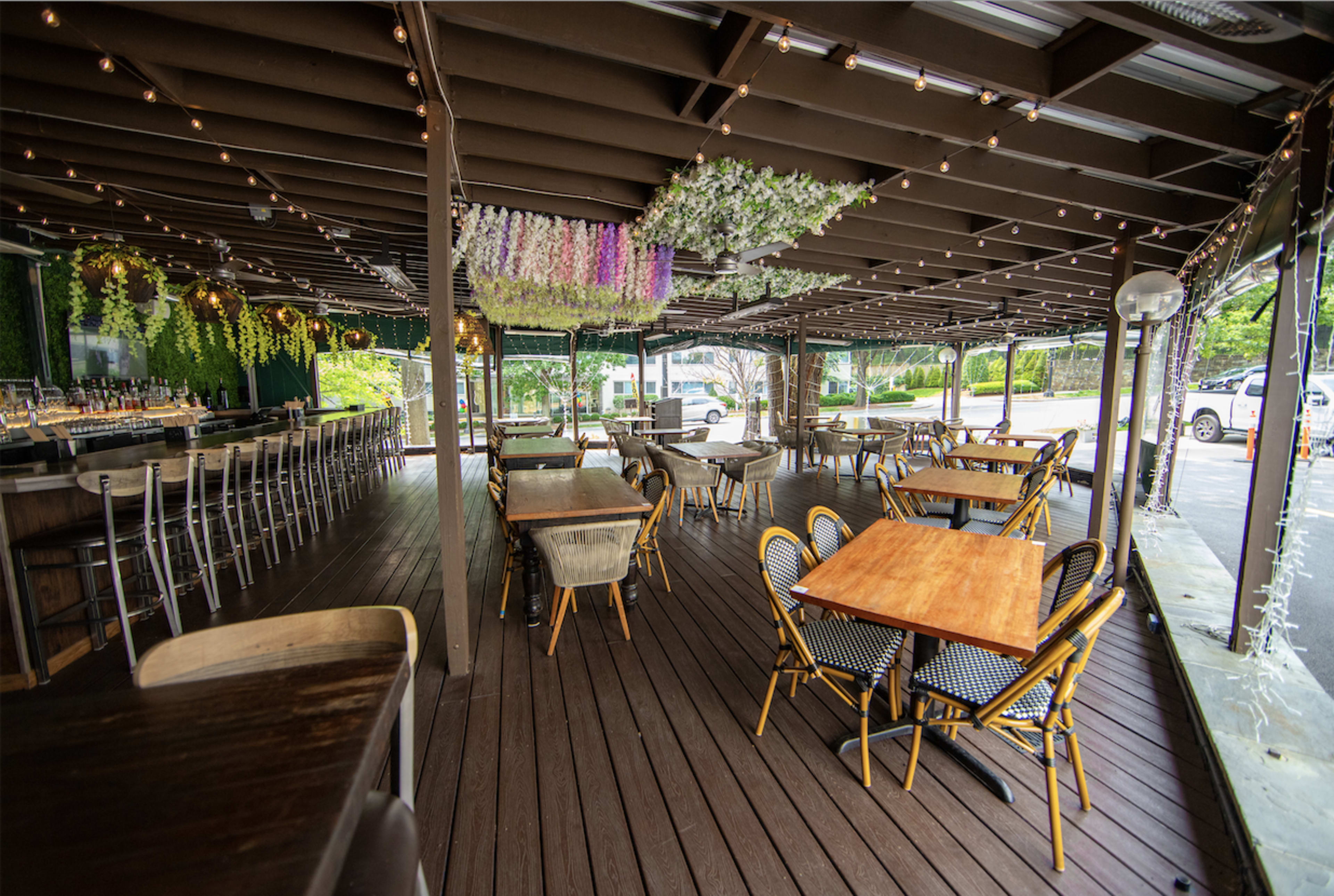 The image shows an outdoor dining area of a restaurant with wooden flooring, tables, and chairs, adorned with hanging floral decorations and string lights.