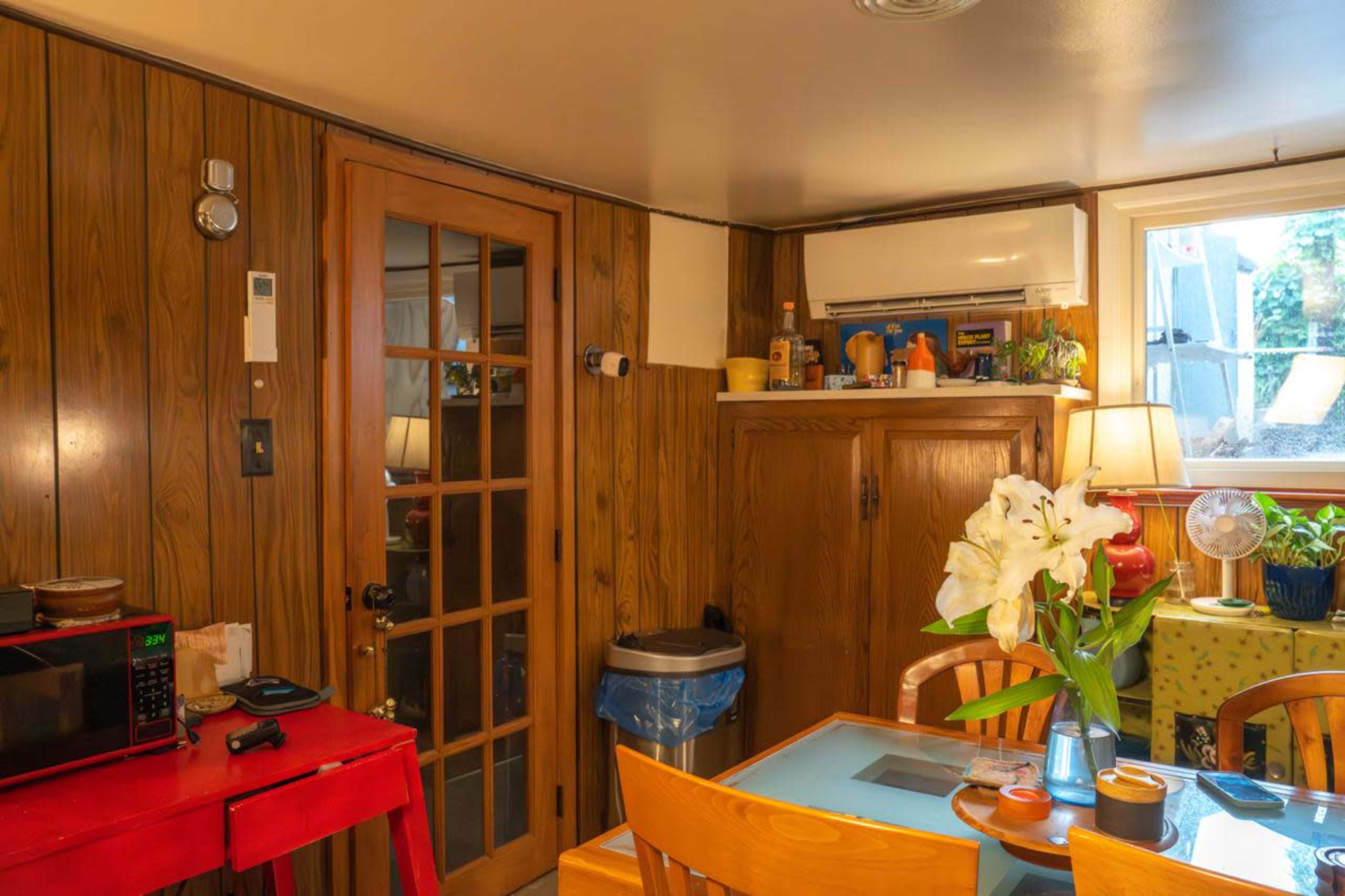 The image shows a cozy kitchen area with wooden paneling, a table with a vase of flowers, a microwave, and a window that lets in natural light.