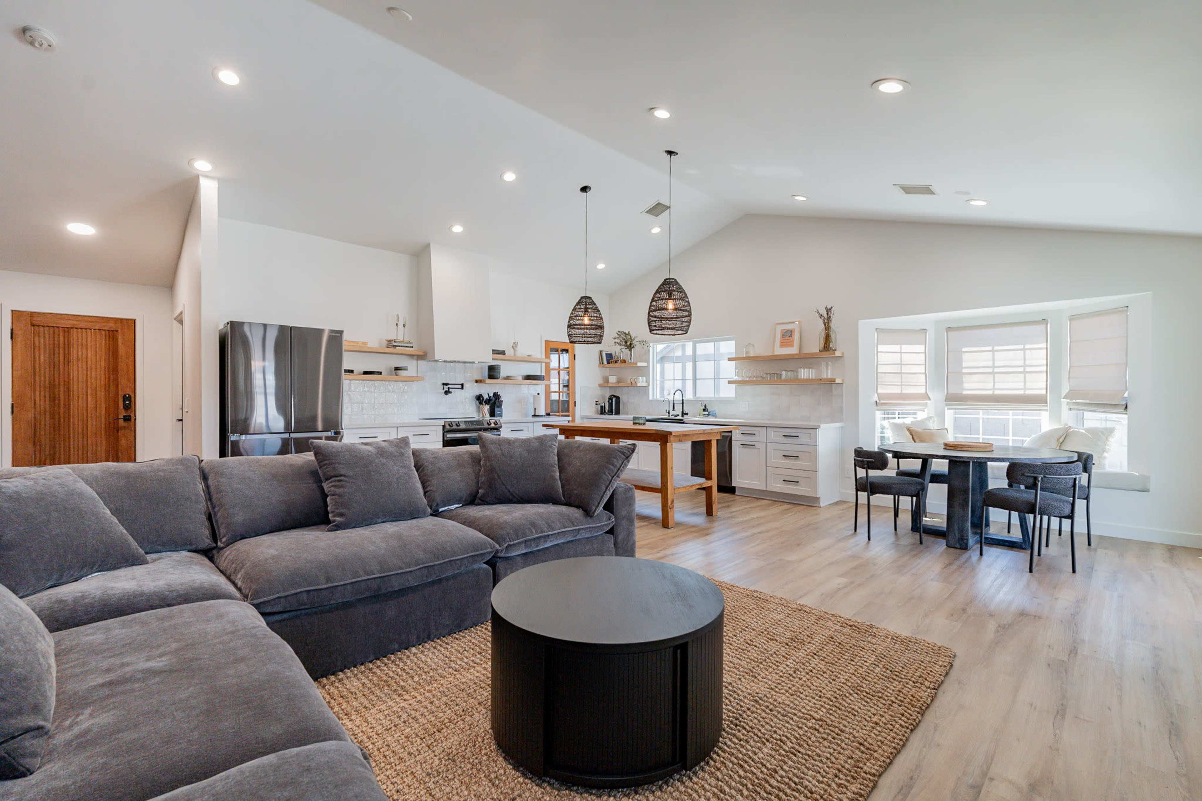 The image shows a modern open-concept living space with a large sectional sofa, a circular coffee table, a kitchen area with sleek cabinetry, and a dining space featuring a small table and chairs.