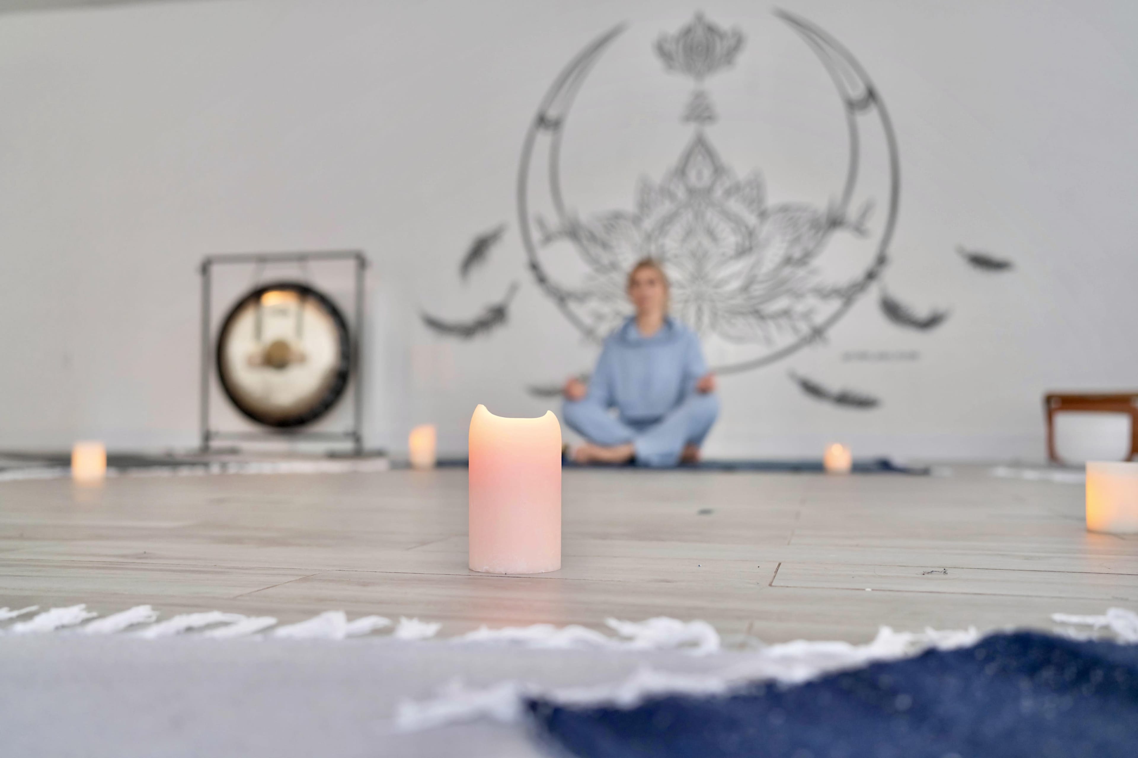 A person sits in a meditative pose on a yoga mat surrounded by lit candles and a wall mural of a lotus flower.
