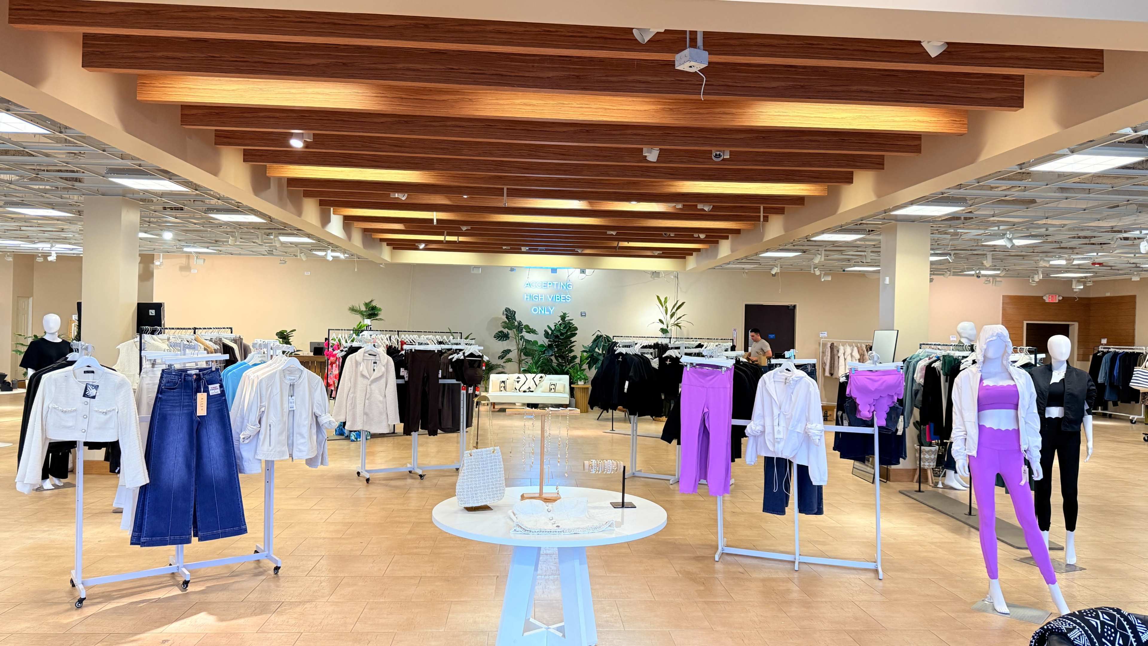 The image shows a spacious retail clothing store with racks of apparel arranged around a central display table, featuring various clothing styles on mannequins and hangers.