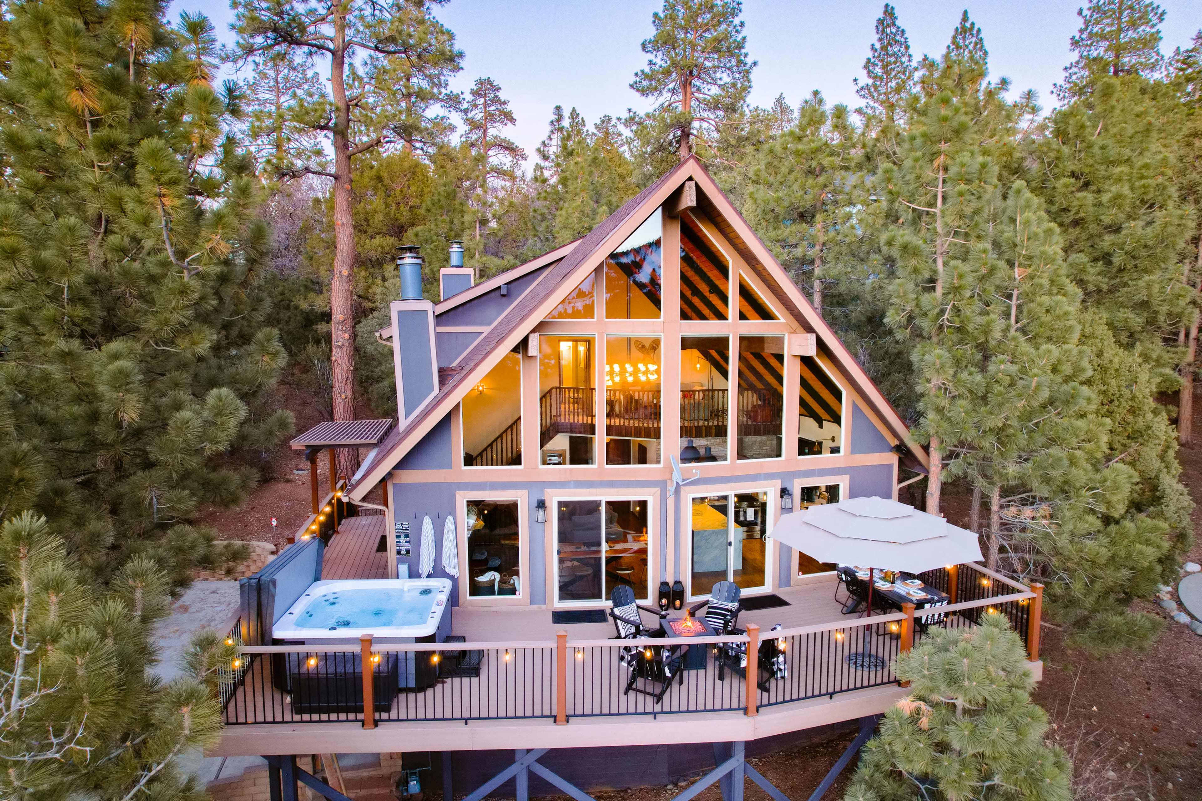 A modern A-frame cabin surrounded by tall pine trees features large windows, a deck with outdoor seating, and a hot tub.