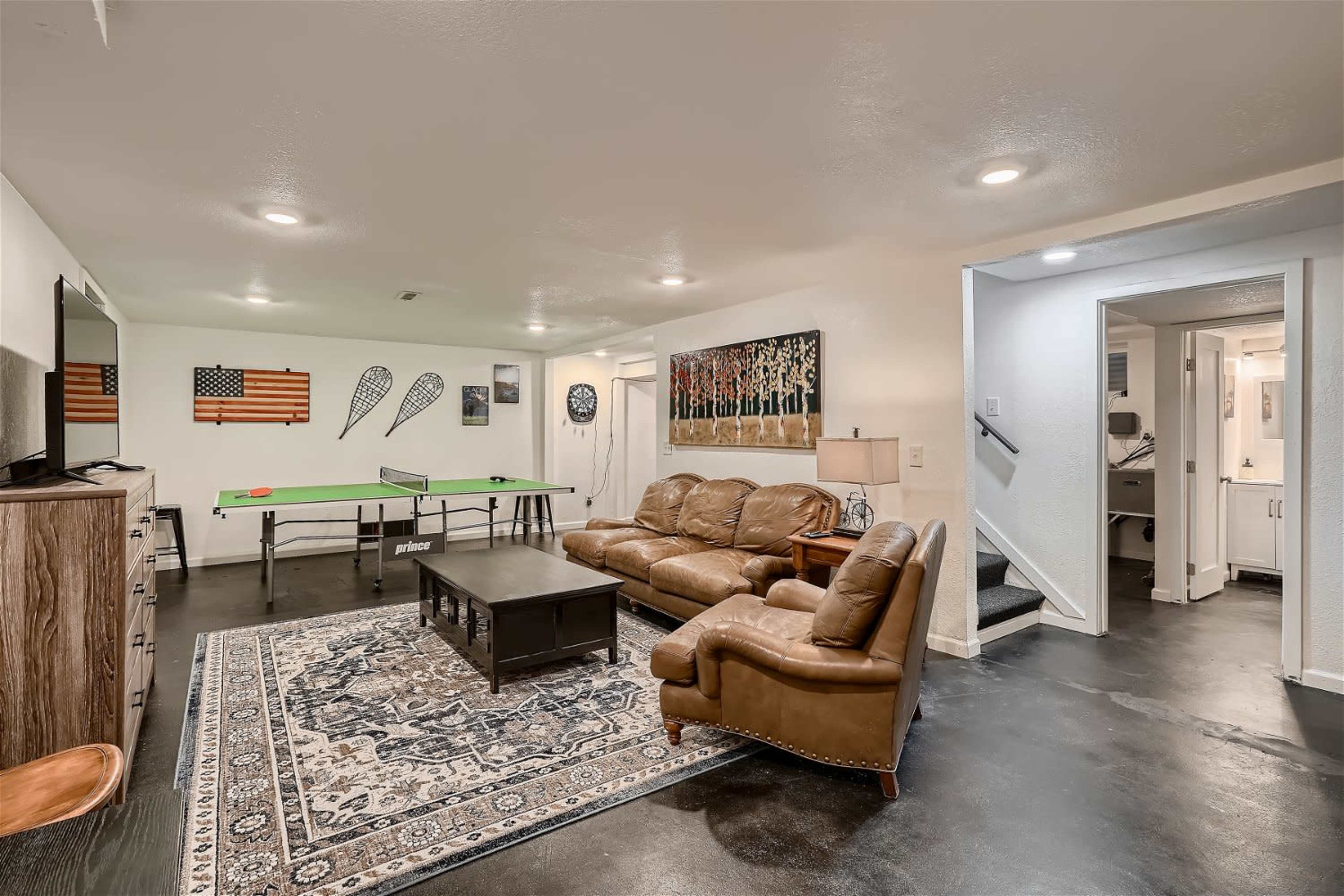The image shows a basement recreation area featuring a ping pong table, a seating arrangement with brown leather couches, and various wall decorations.