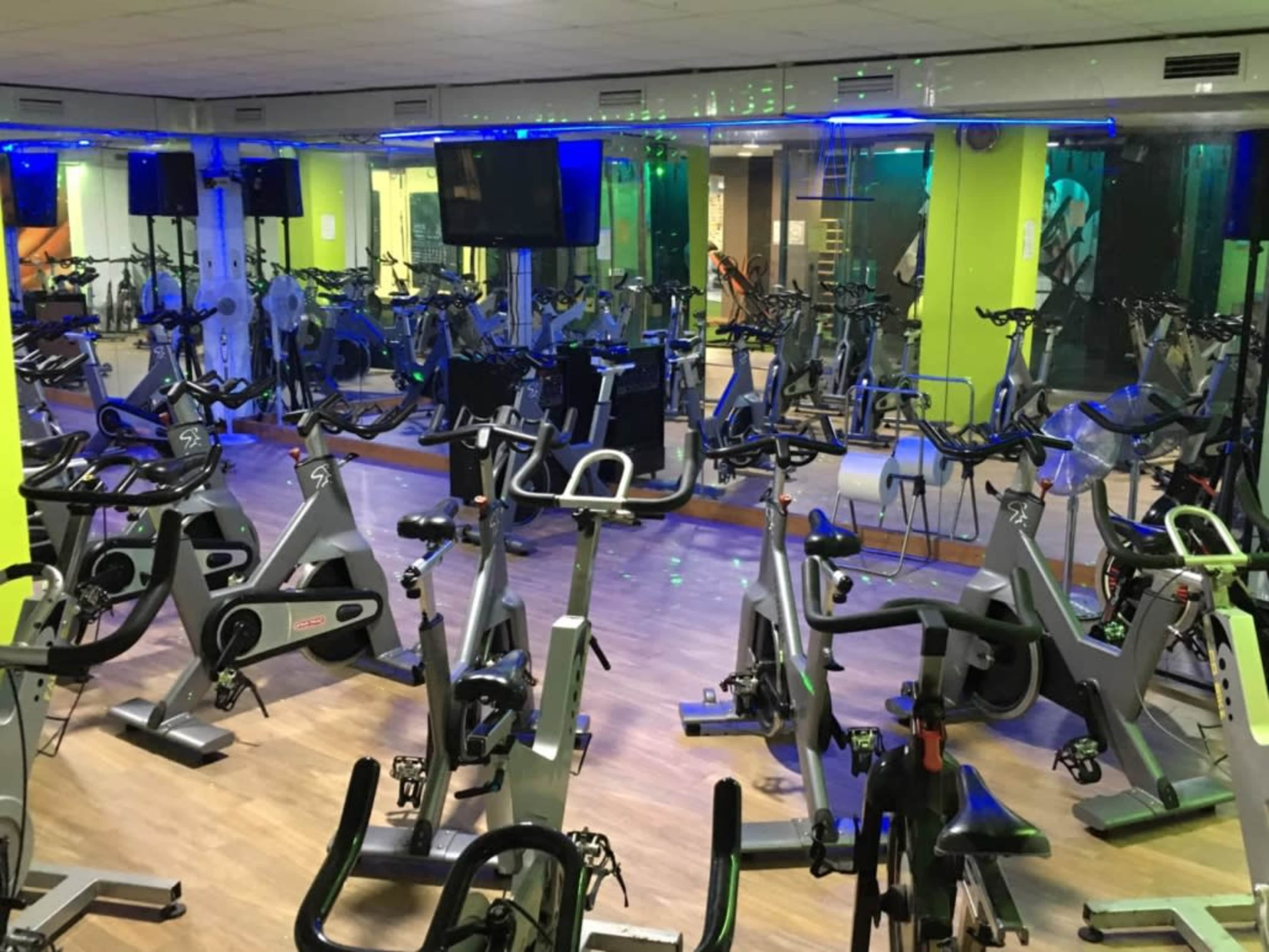 The image shows a spacious indoor cycling studio with multiple exercise bikes arranged in rows and vibrant lighting.