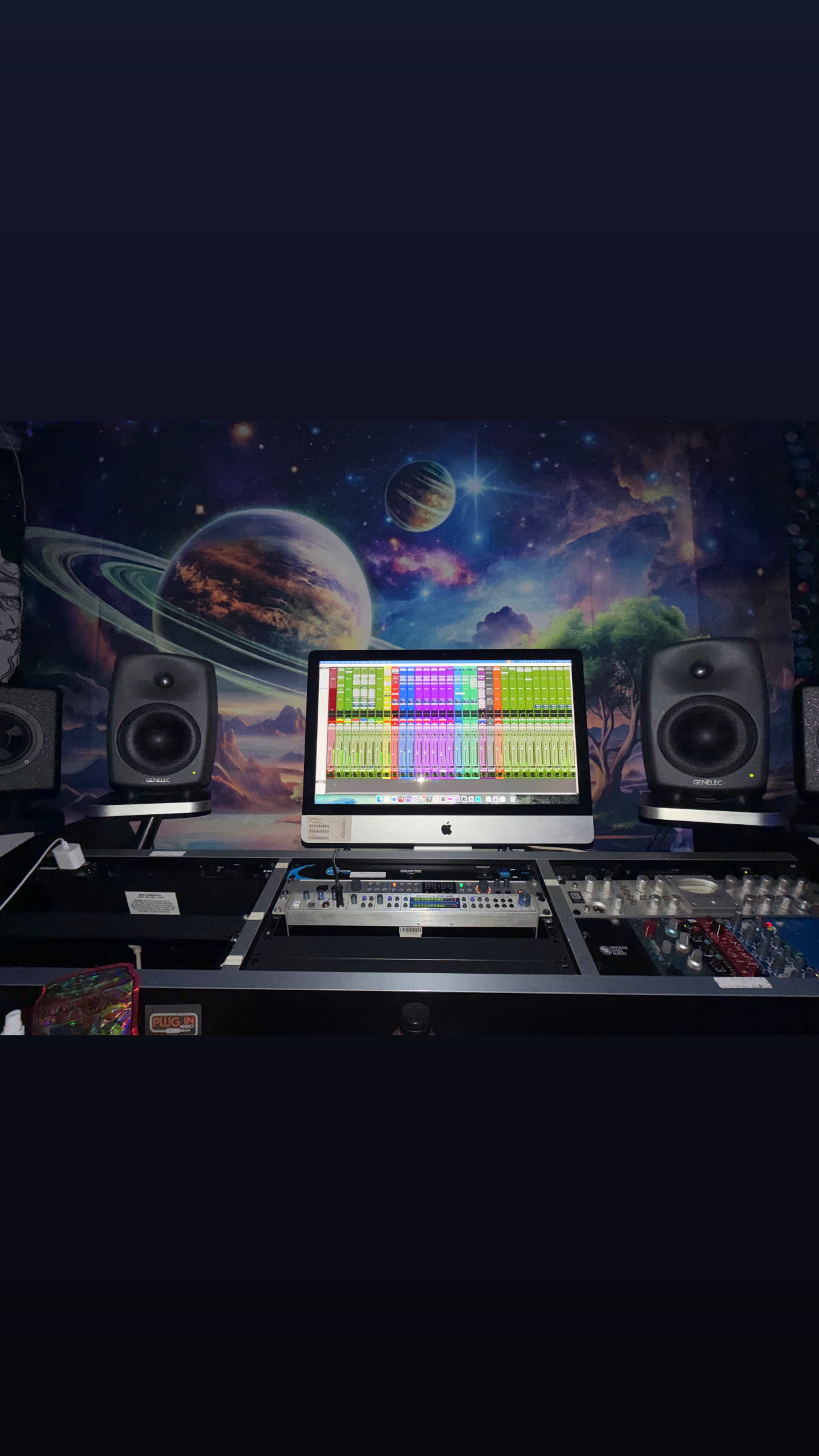 The image shows a music production setup featuring an iMac monitor displaying audio software, flanked by two speakers, against a backdrop of a space-themed mural.