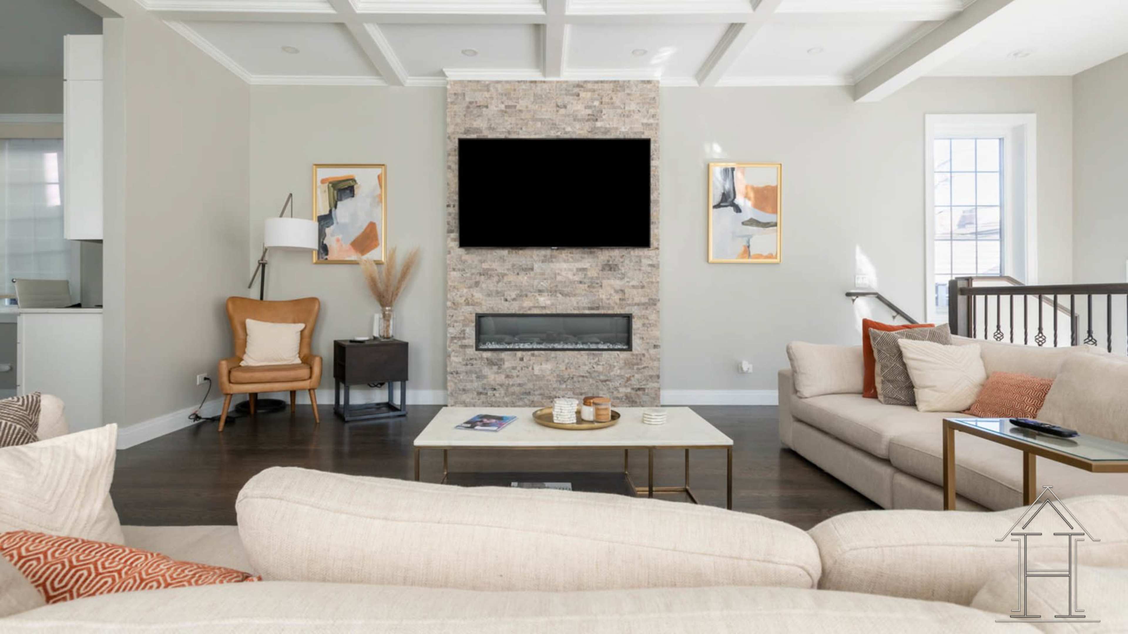 A modern living room features a stone fireplace with a large blank screen above it, flanked by artwork, and furnished with a beige sofa and a cozy armchair.