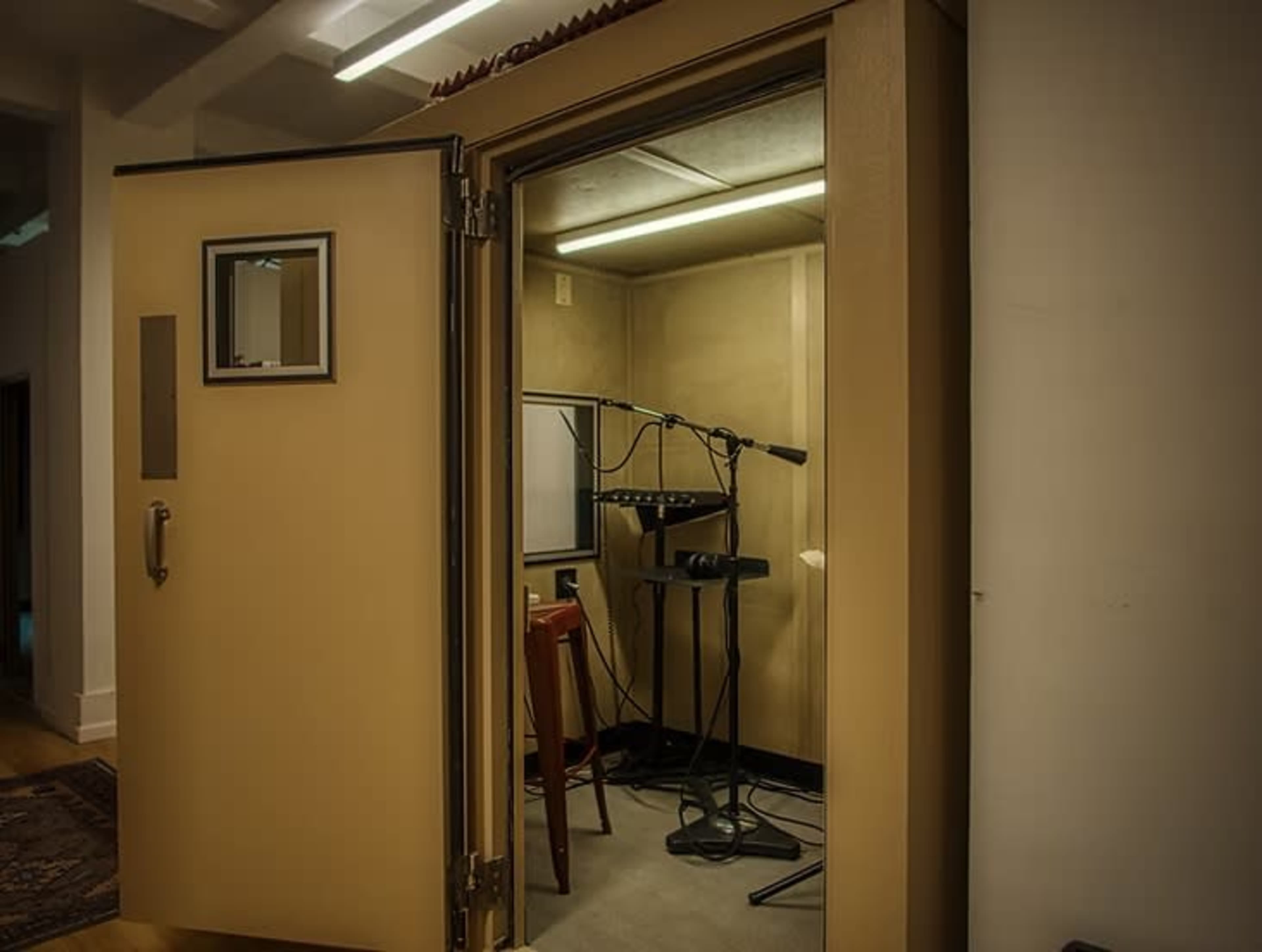 The image shows an open sound booth with a microphone setup and a small desk inside.
