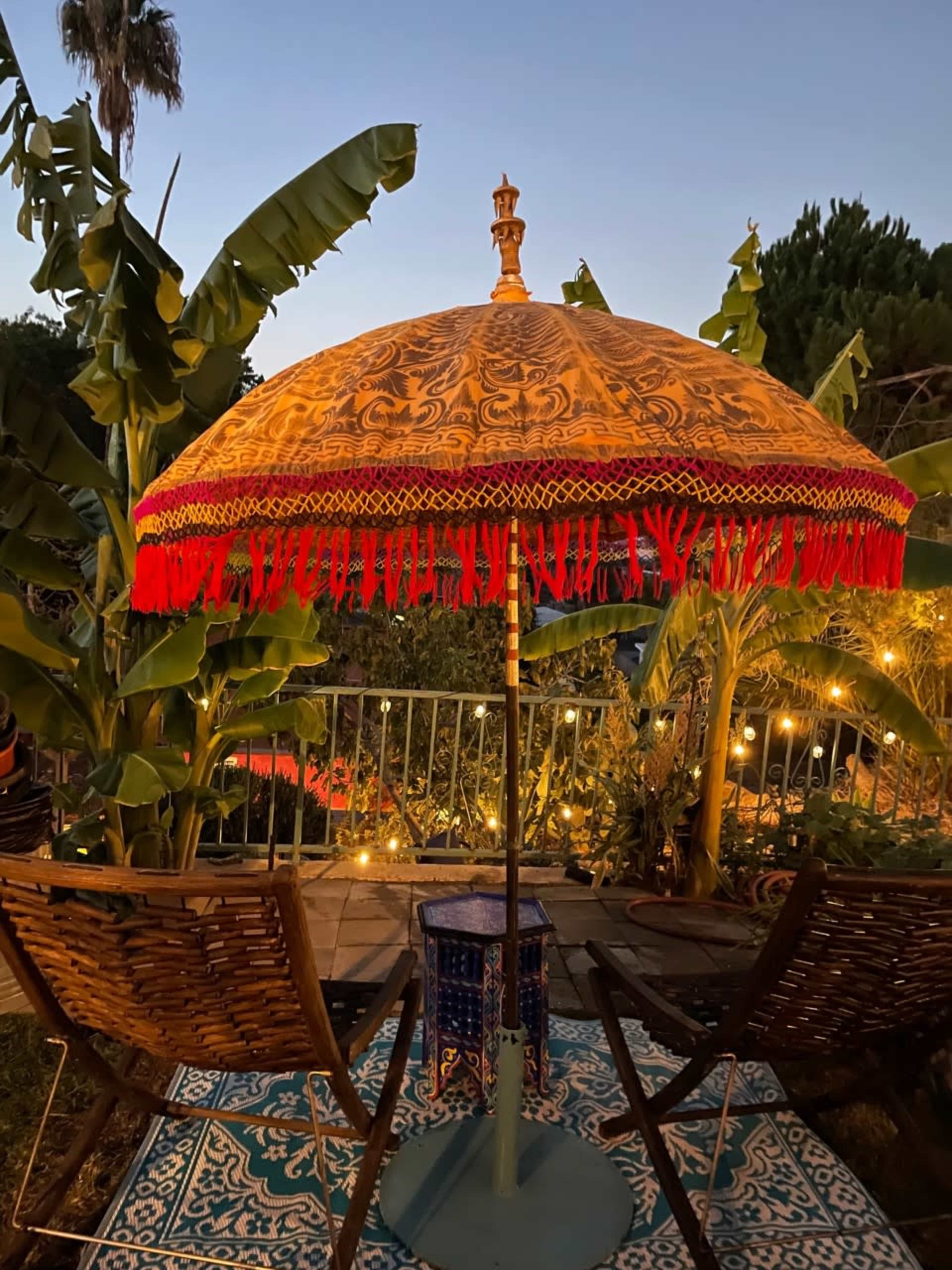 A decorative umbrella stands between two wooden chairs on a patterned rug, surrounded by lush greenery and soft evening lights.