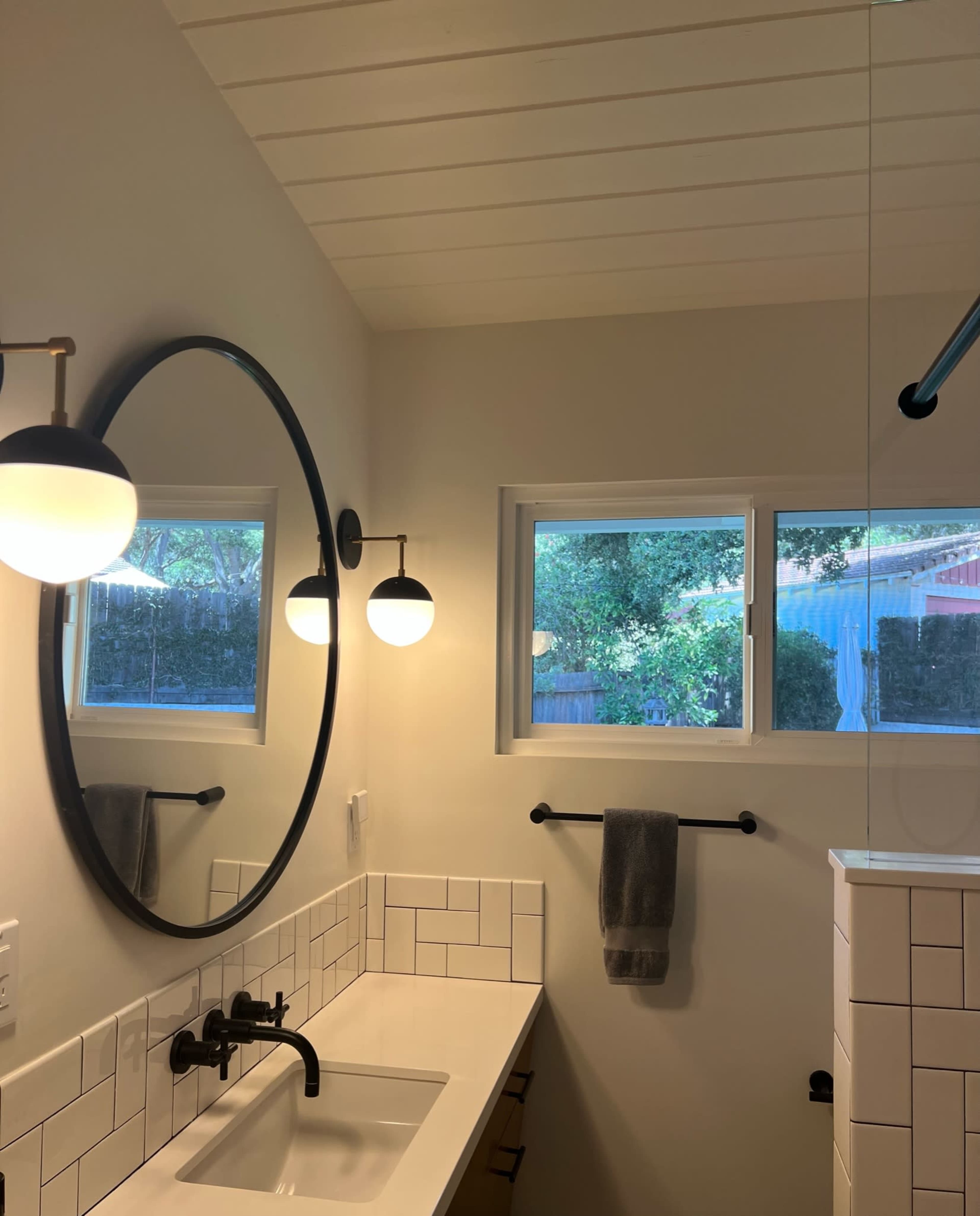 A bathroom features a large round mirror above a white sink, with two pendant lights and a gray towel hanging on a wall bar, illuminated by natural light from windows.