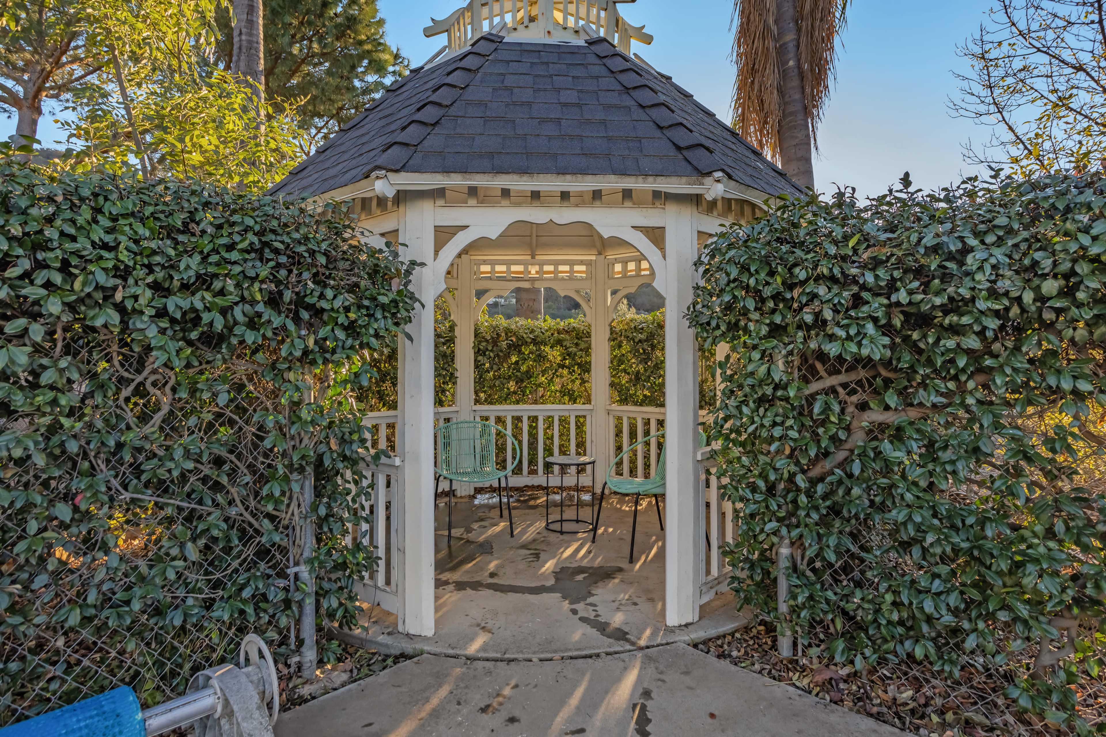 A white gazebo with a shingled roof is surrounded by lush greenery and features two chairs inside.