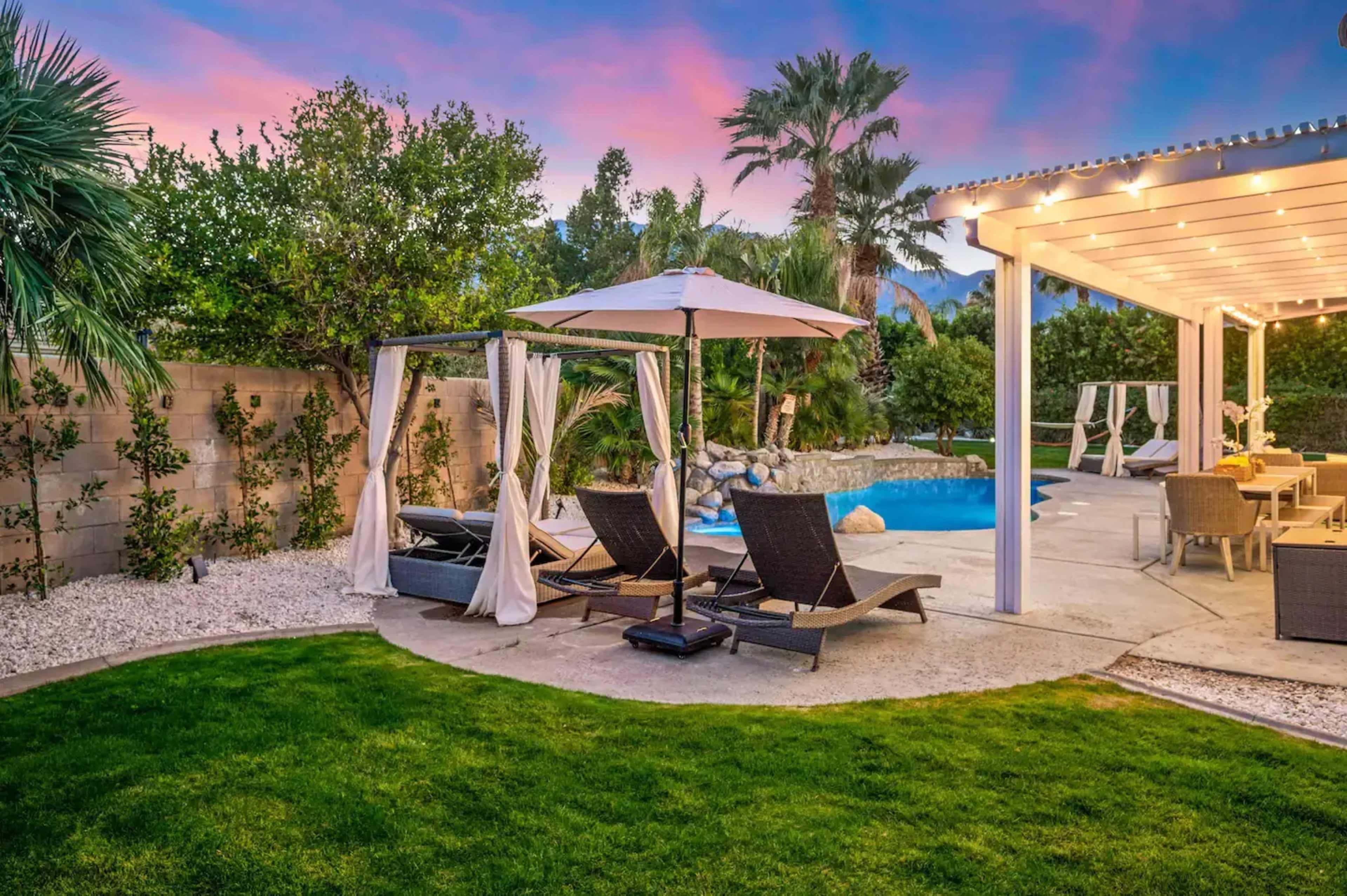 The image shows a landscaped backyard featuring a swimming pool, lounge chairs with an umbrella, and a covered seating area under string lights.