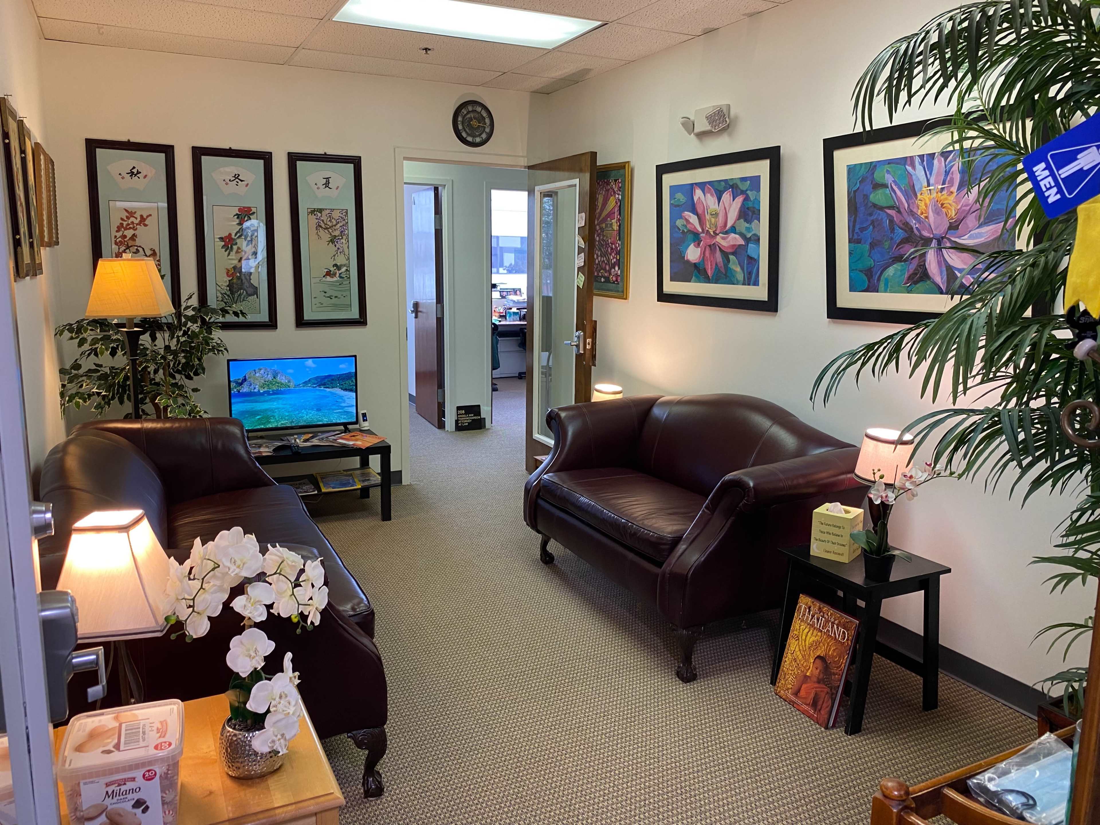 The image shows a cozy office waiting area furnished with two leather sofas, a television, decorative artwork on the walls, and potted plants.