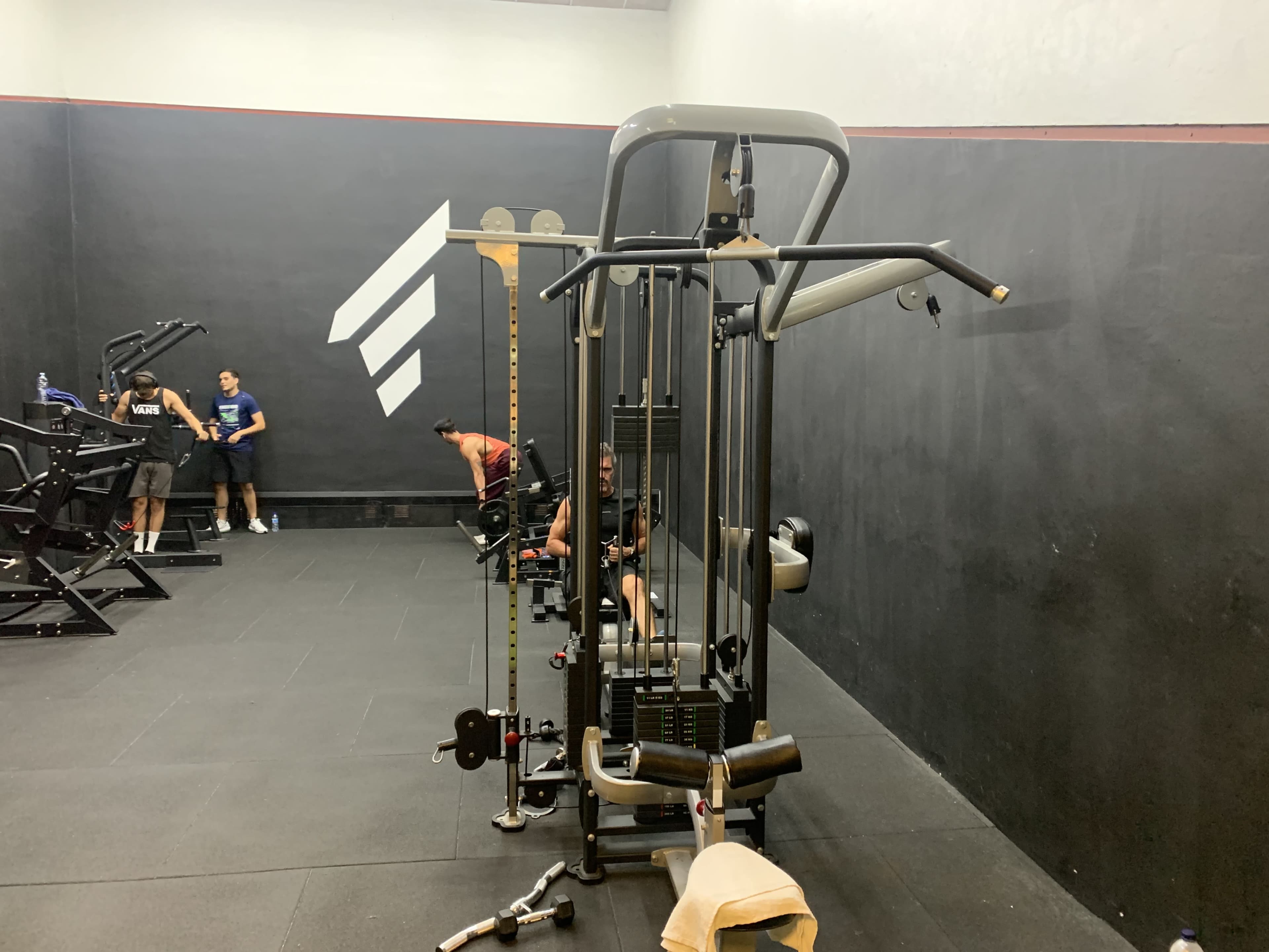 A gym interior with various exercise equipment, including a cable machine, and individuals engaging in workouts.
