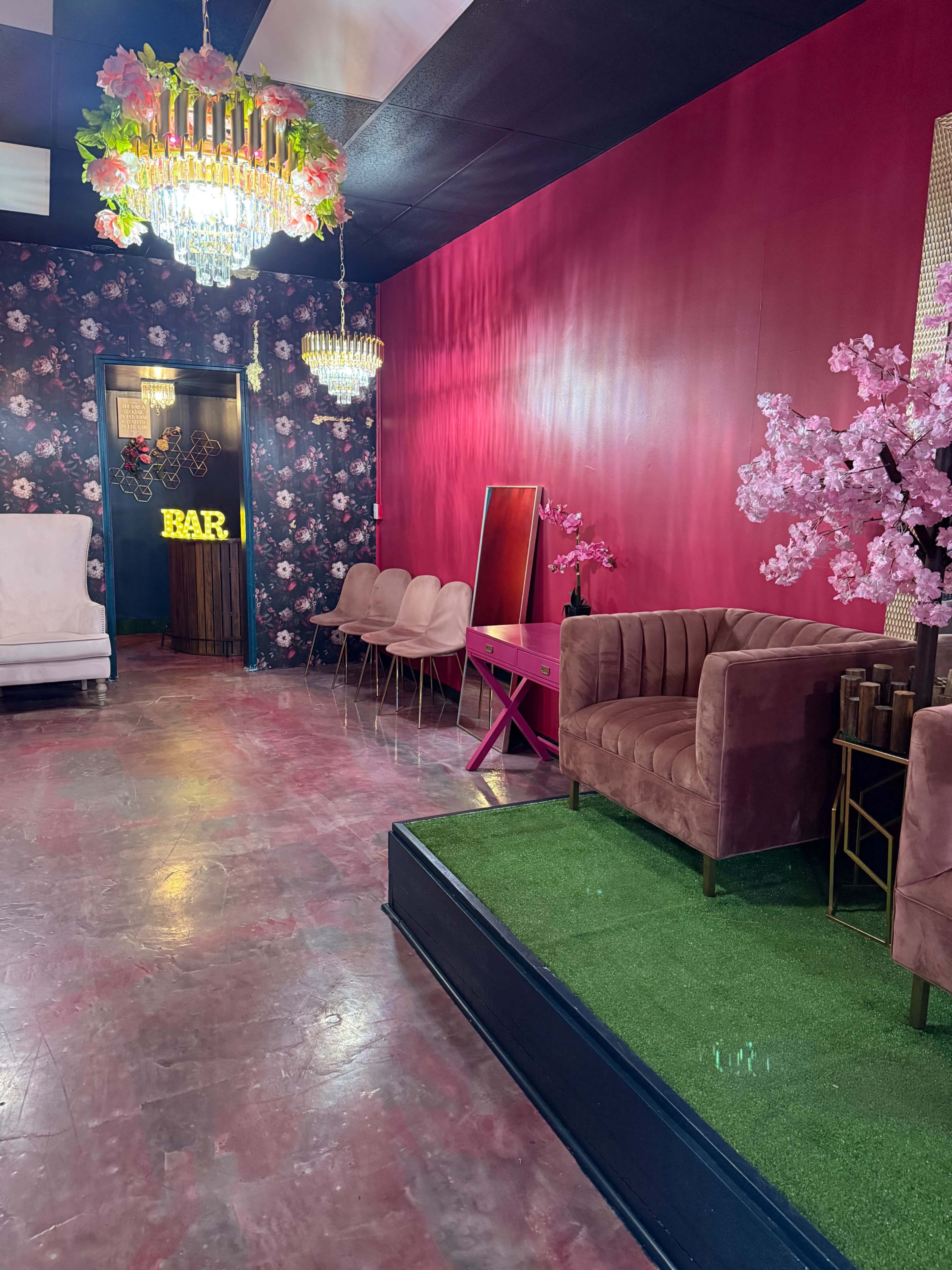 The image shows a vibrant interior space featuring pink walls, floral wallpaper, a chandelier, and modern seating arrangements.