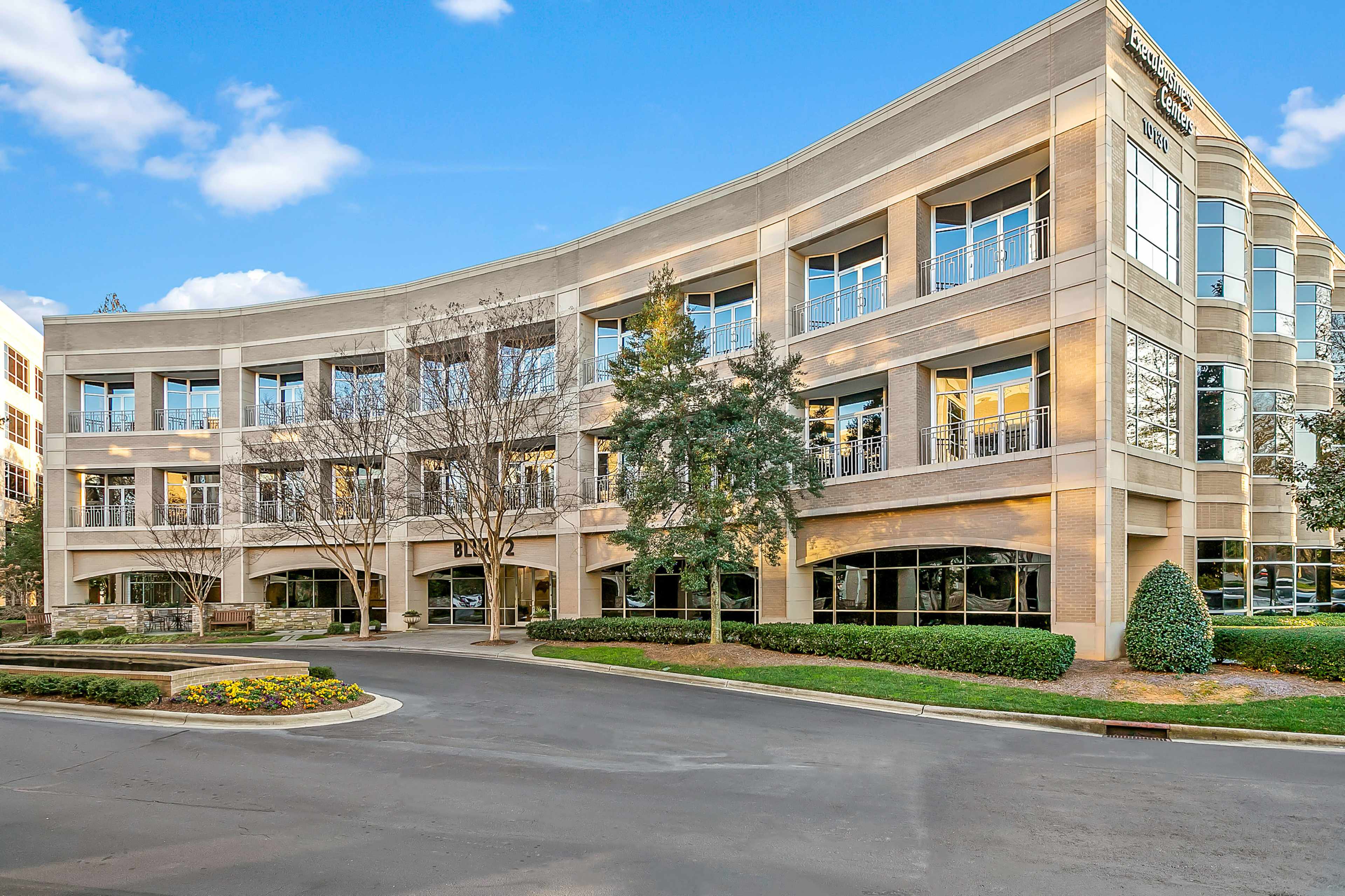 A modern, multi-story office building with large windows and a curved facade, set in a landscaped area with greenery and parking space.