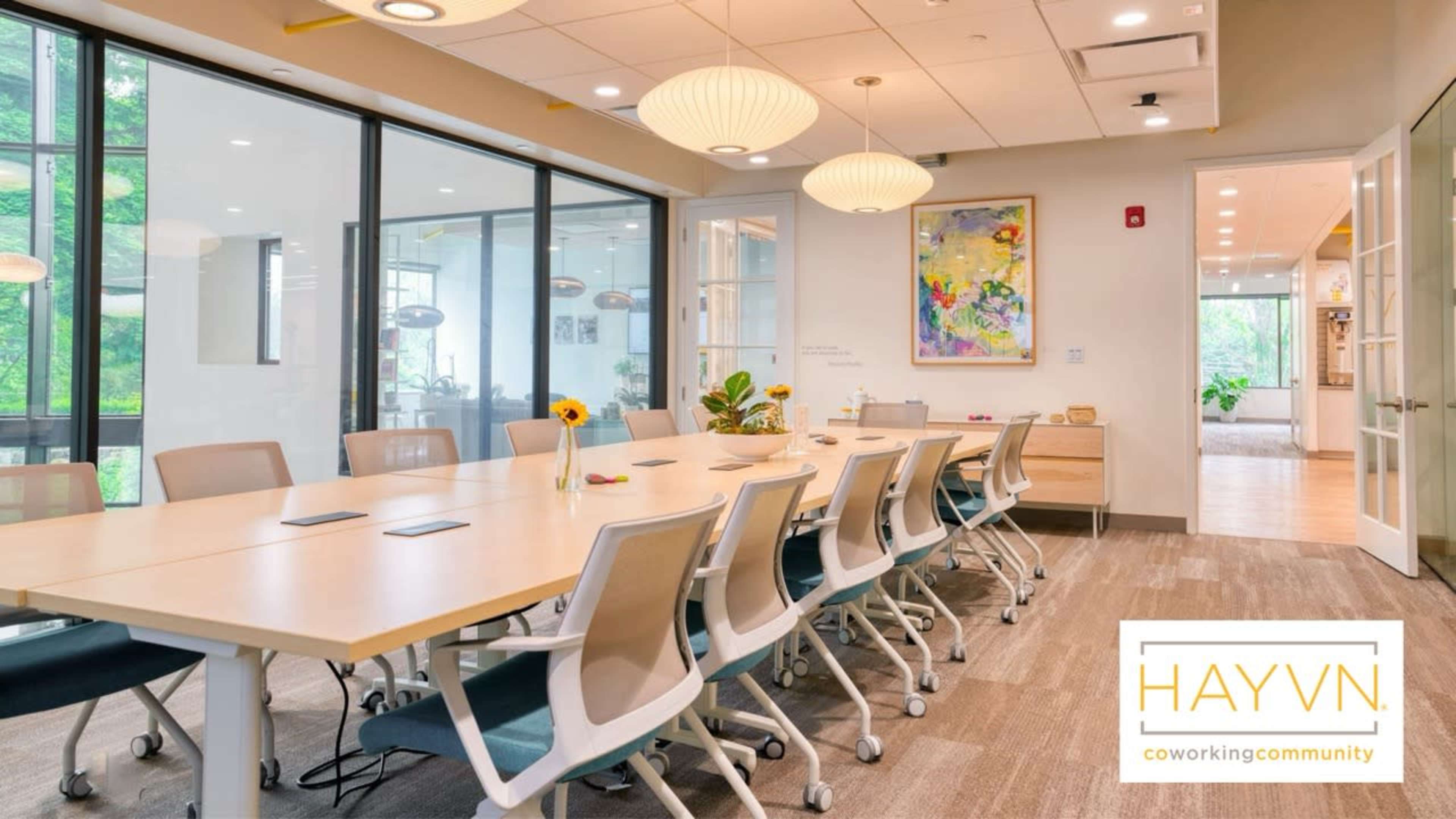 A large conference room features a long wooden table surrounded by white chairs, with large windows allowing natural light to illuminate the space.