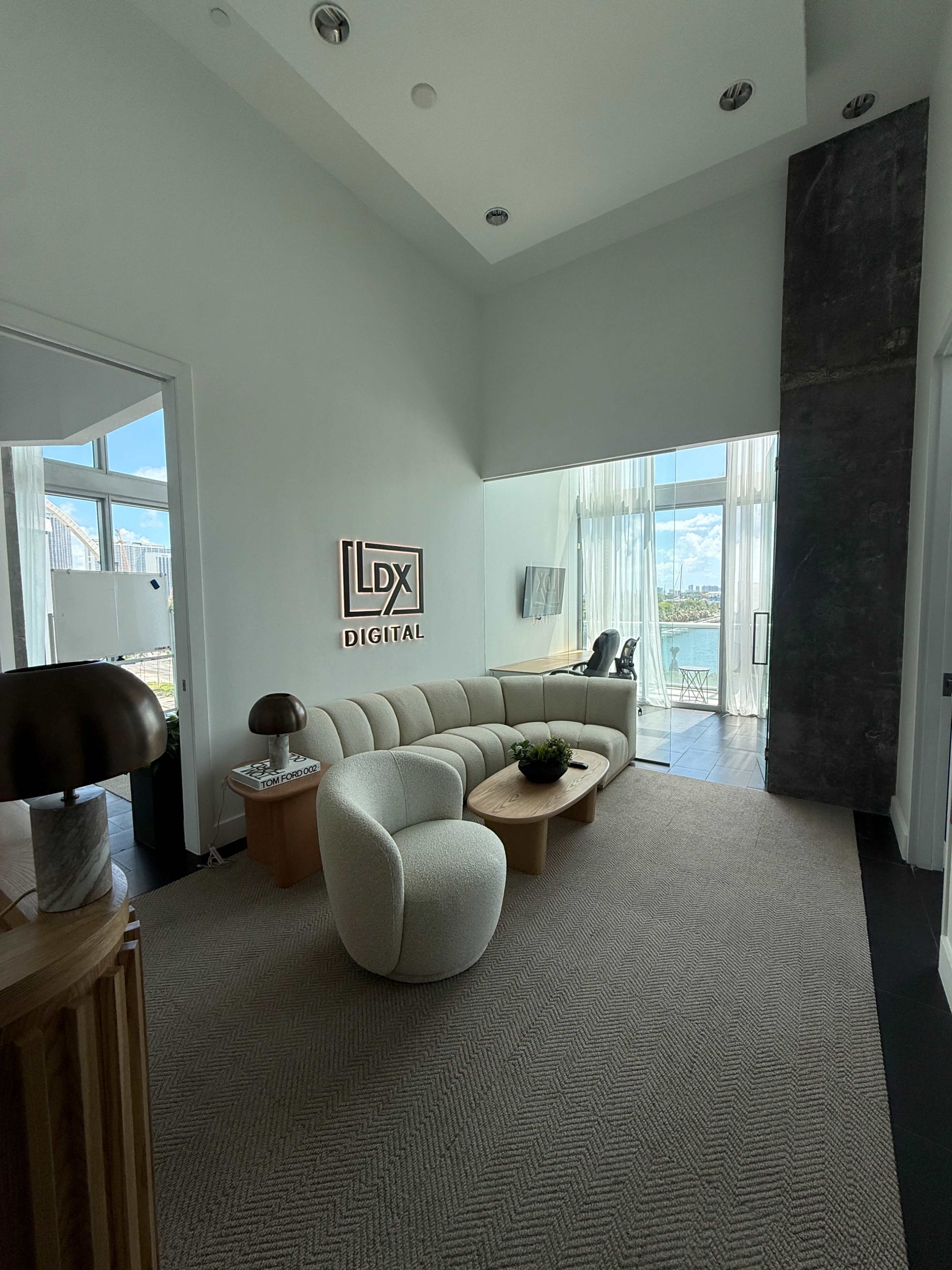 The image shows a modern office space with a light-colored curved sofa, a round wooden coffee table, and a sign that reads "LDX Digital" on the wall, alongside large windows that let in natural light.