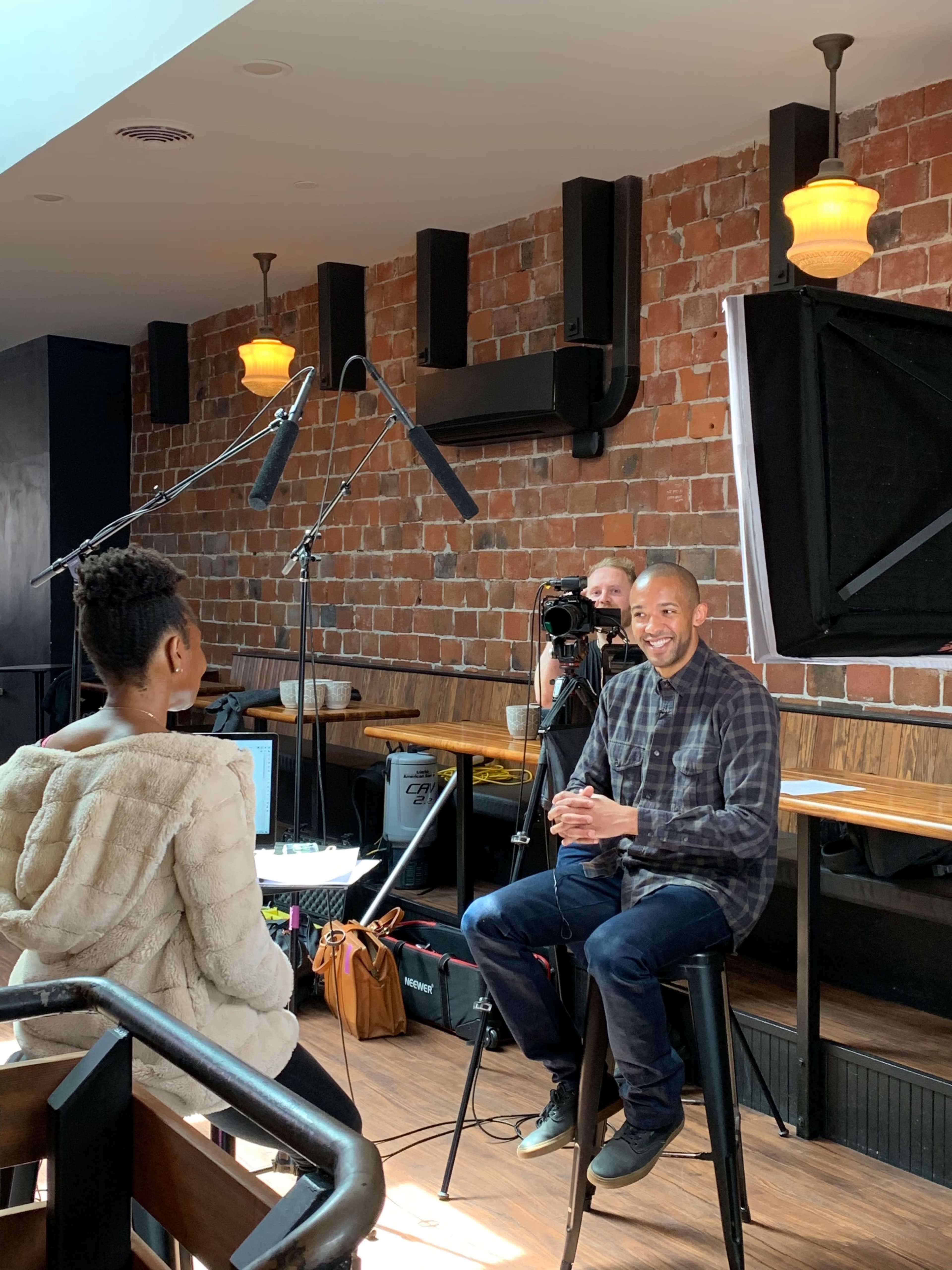 A man in a plaid shirt sits on a stool while being filmed in a restaurant with a woman facing him.