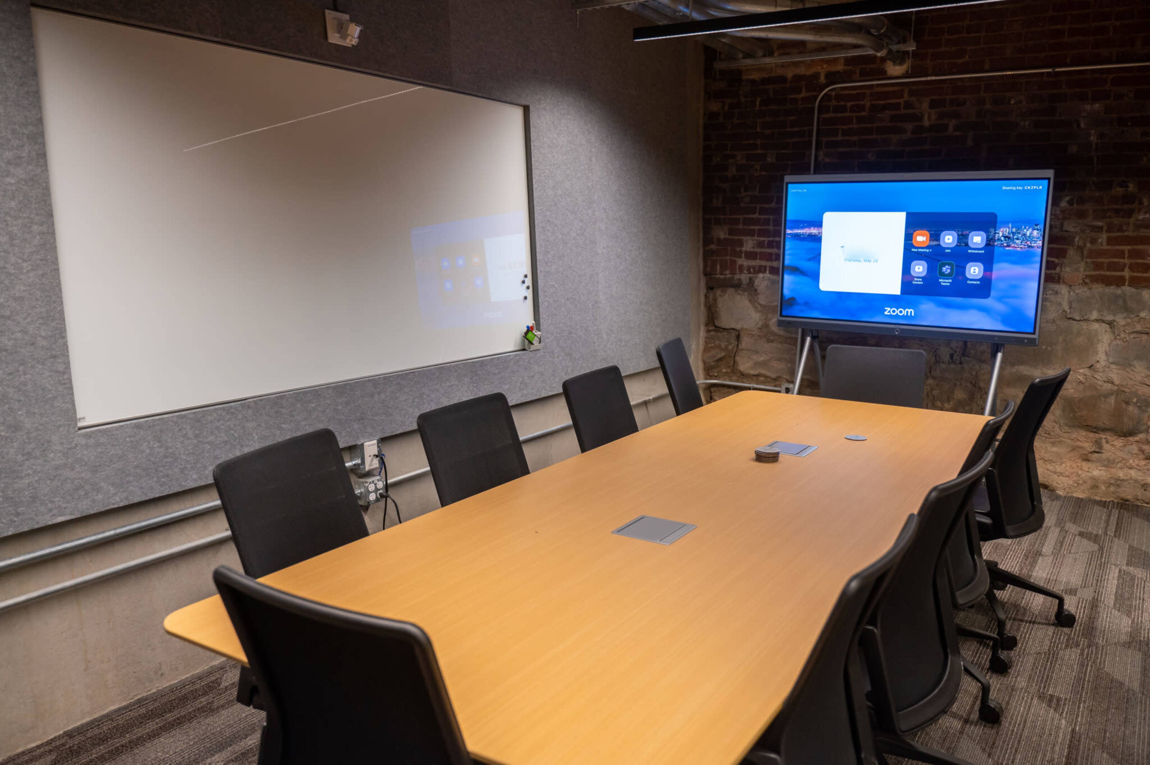 A conference room features a long wooden table surrounded by black chairs, with a large screen displaying a Zoom interface and a blank whiteboard on the wall.