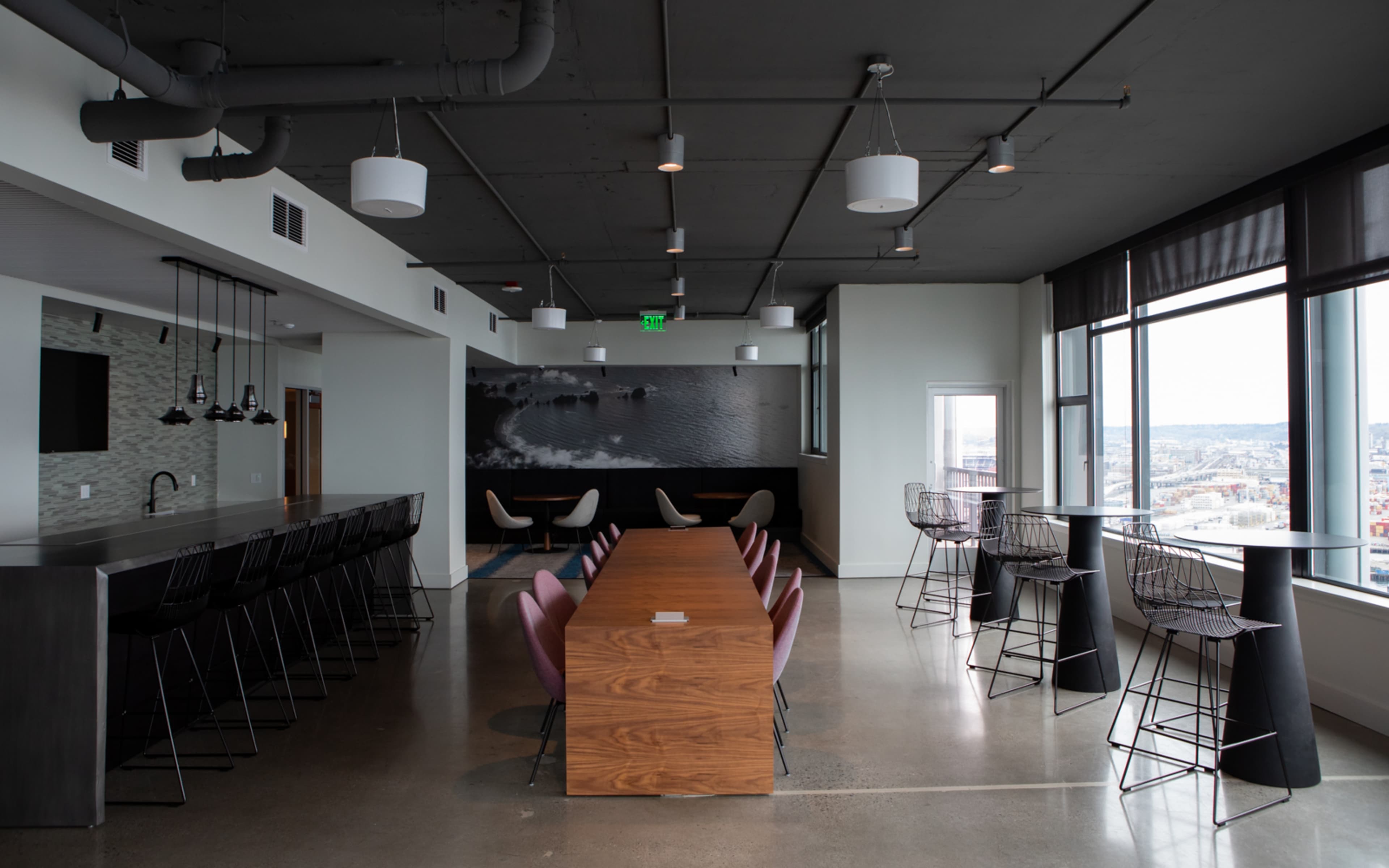 A modern office space featuring a long wooden table, several high chairs, and large windows with a city view.