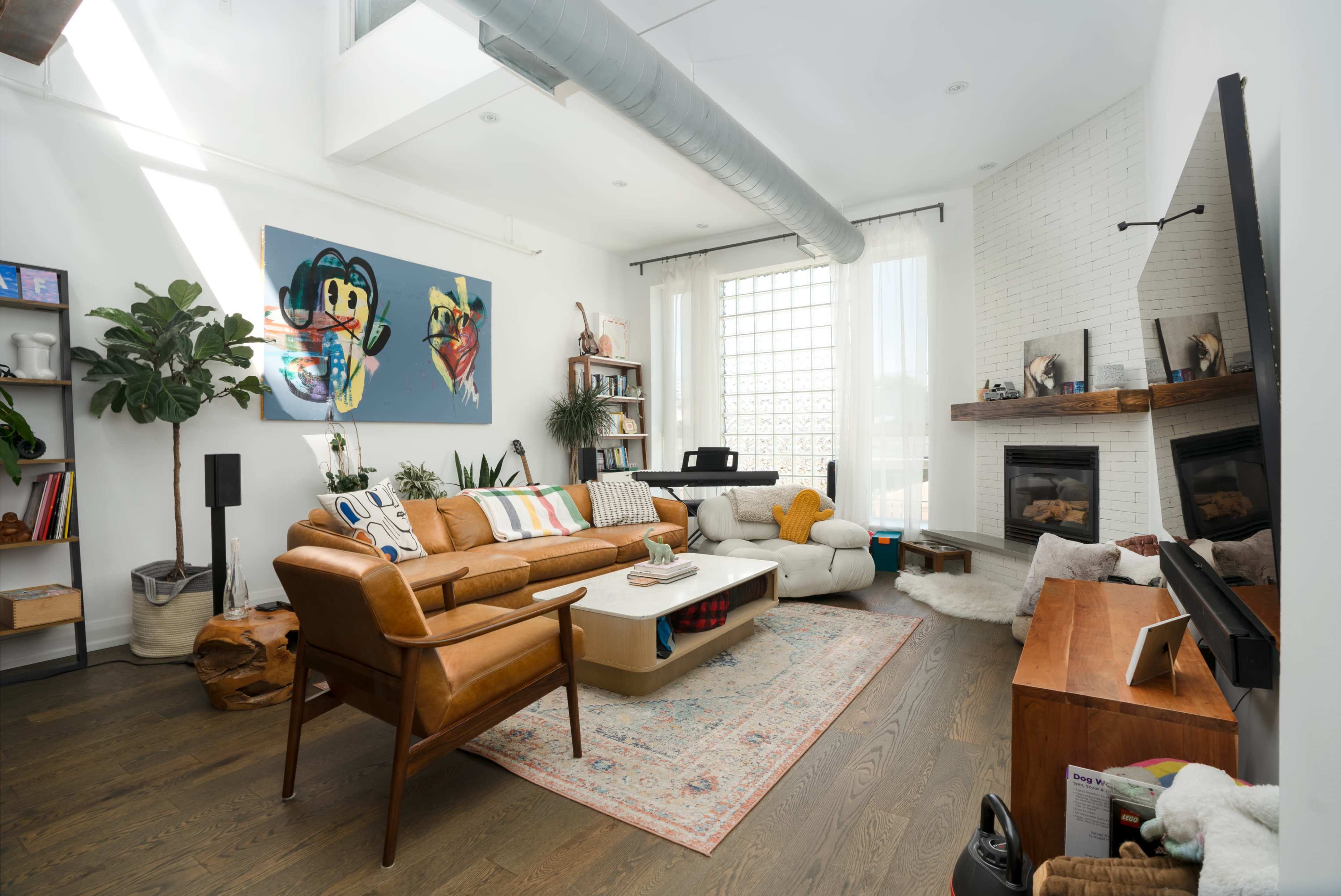 The living room features a brown leather sofa, a white coffee table, a fireplace, and large windows, with artwork and plants adorning the walls.