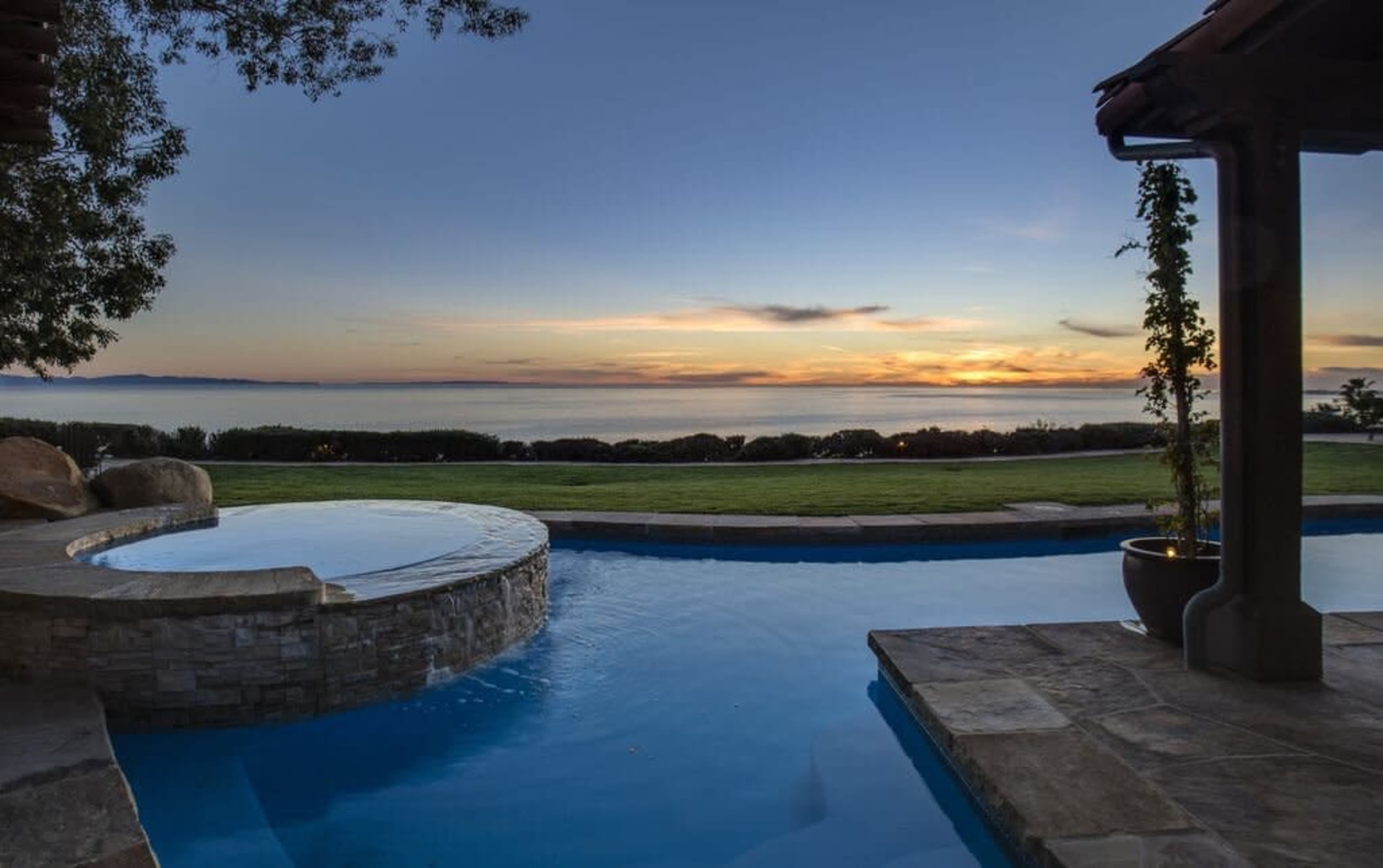 A serene poolside view featuring a circular hot tub, lush green grass, and a sunset over the water in the background.