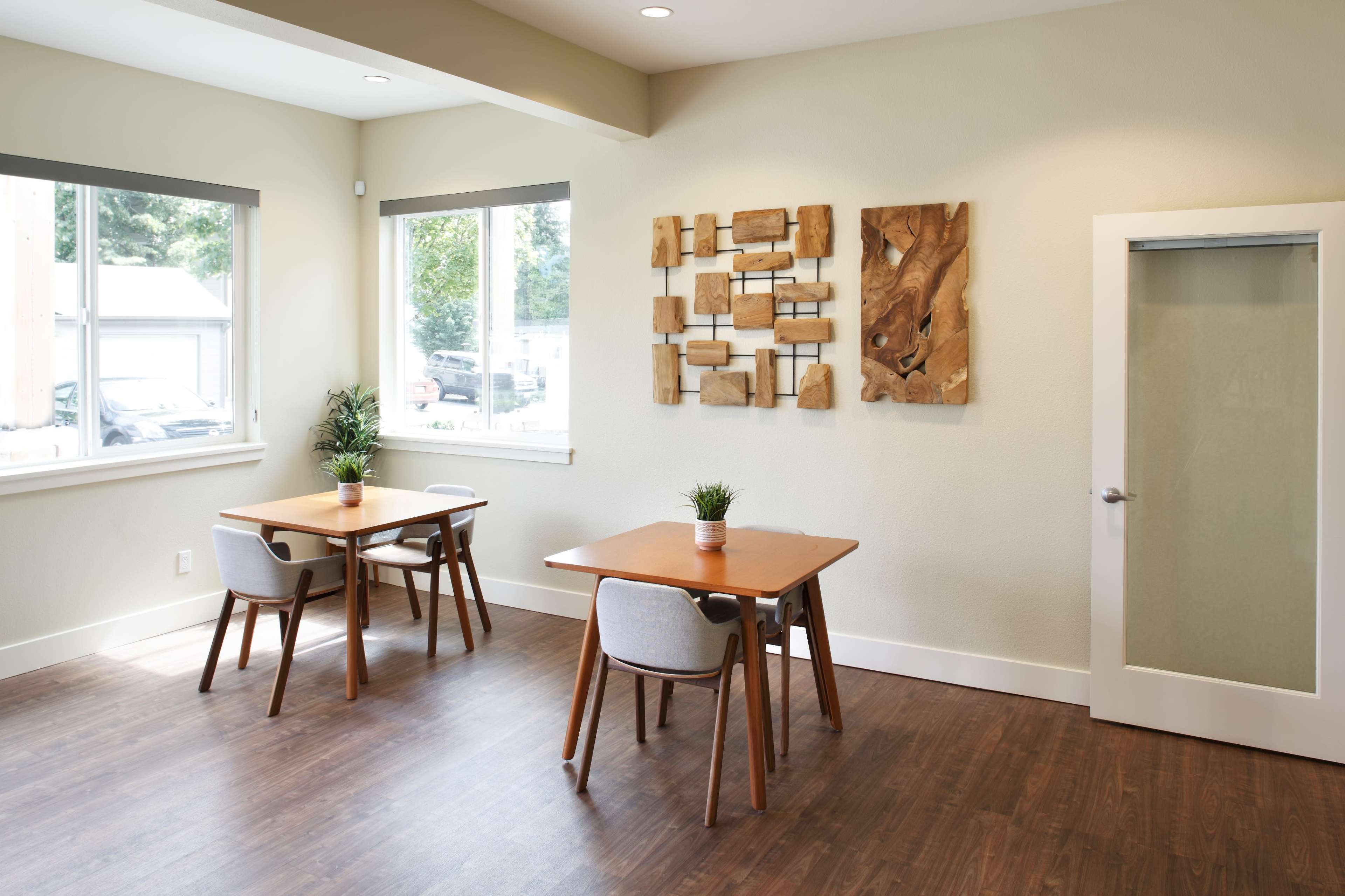 A light-colored room with two wooden tables and chairs, decorated with a wooden wall art piece, and a glass-paneled door.