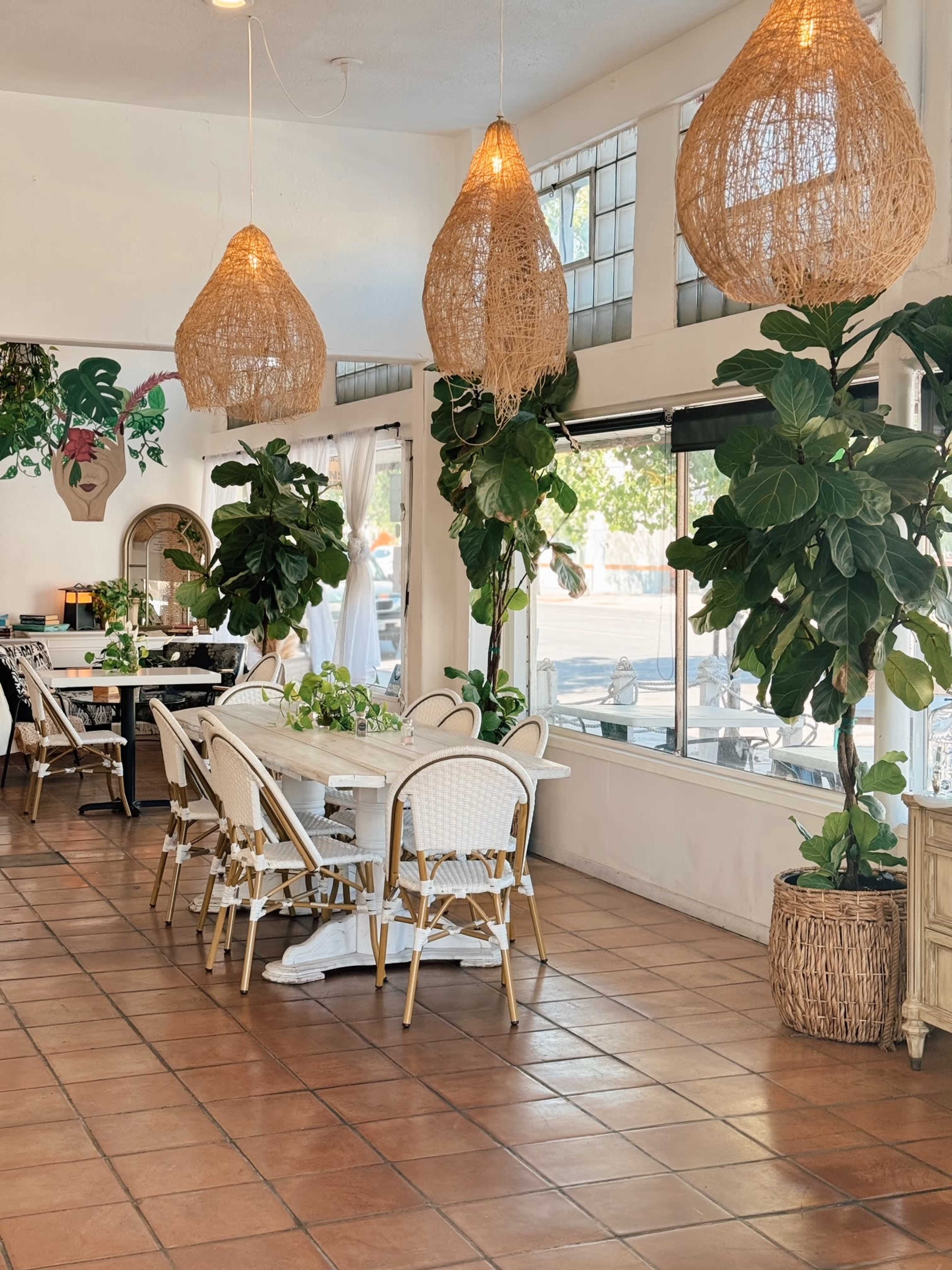 A dining area with a large white table surrounded by wicker chairs, illuminated by three woven pendant lights, and decorated with potted plants.