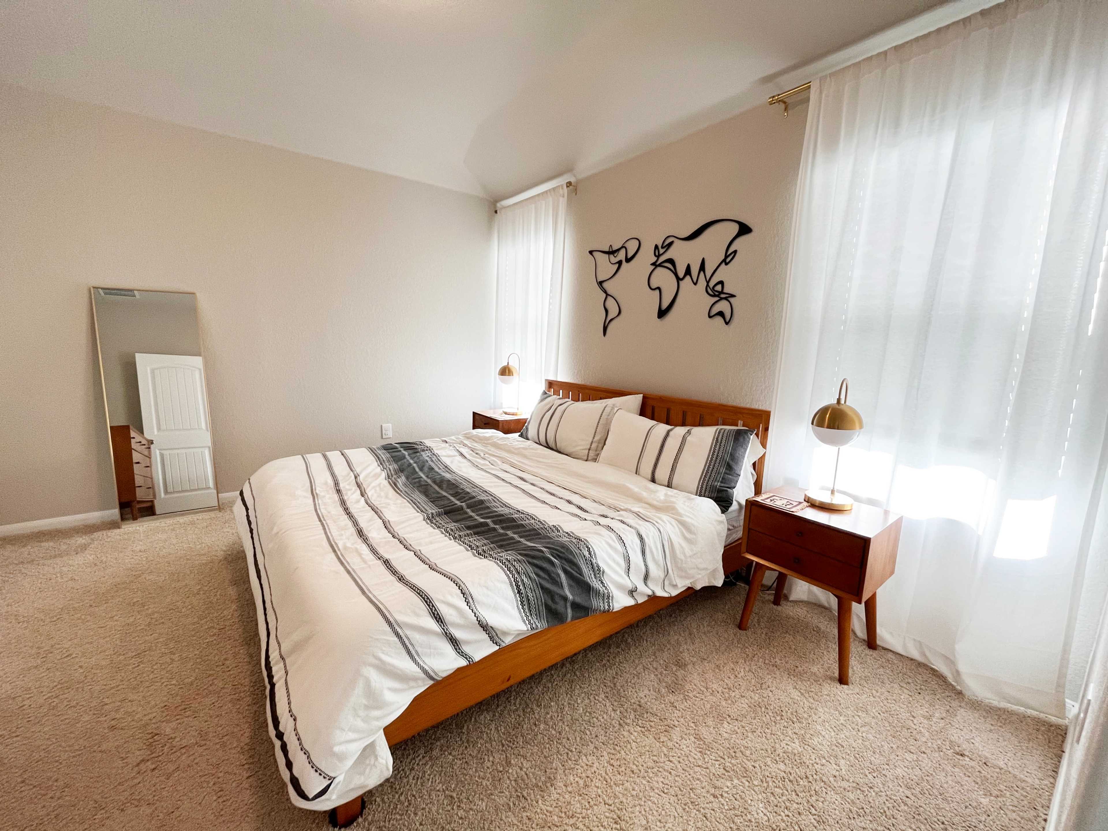 A neatly arranged bedroom features a wooden bed with a striped duvet, two bedside tables with lamps, and large windows dressed with sheer curtains.